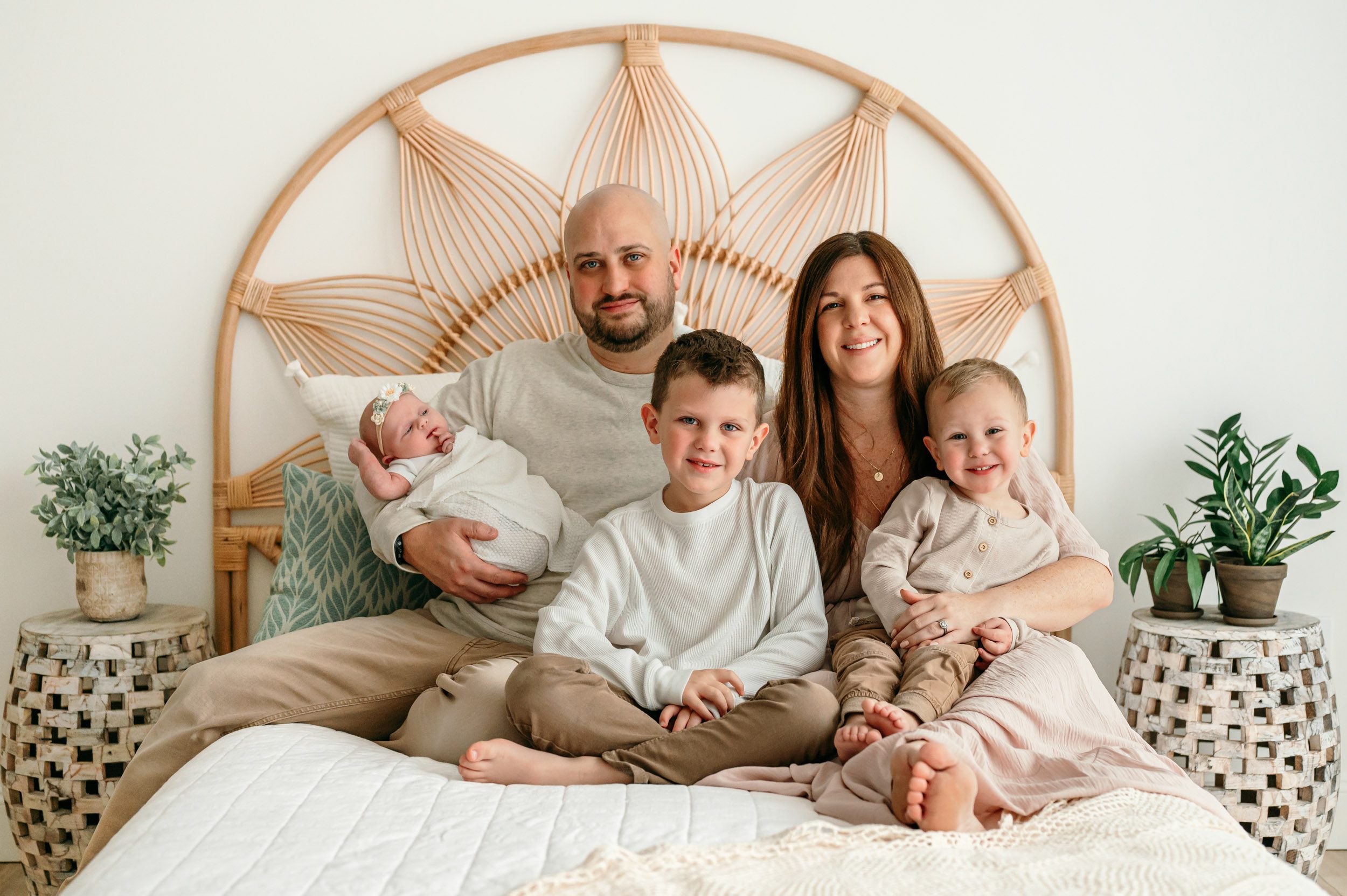 a mom and dad sitting on a bed with their baby girl and two young sons sitting on their laps as they all smile at the camera during a natural light newborn photoshoot