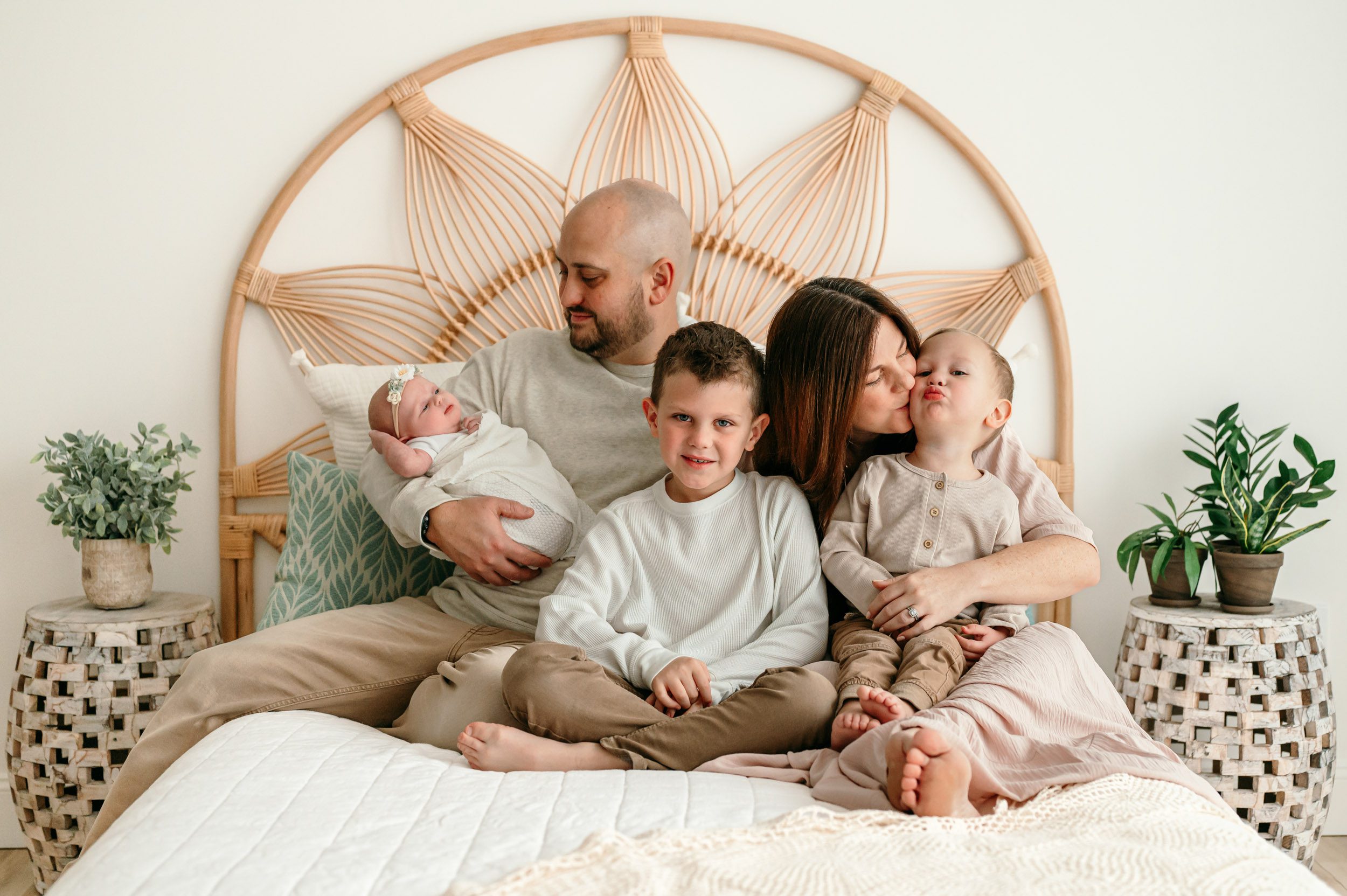 a family of five sitting on a bed snuggling together while dad holds his baby girl in his arms and smiles down at her, mom kisses her younger son on the cheek, and big brother smiles at the camera during a newborn photo session