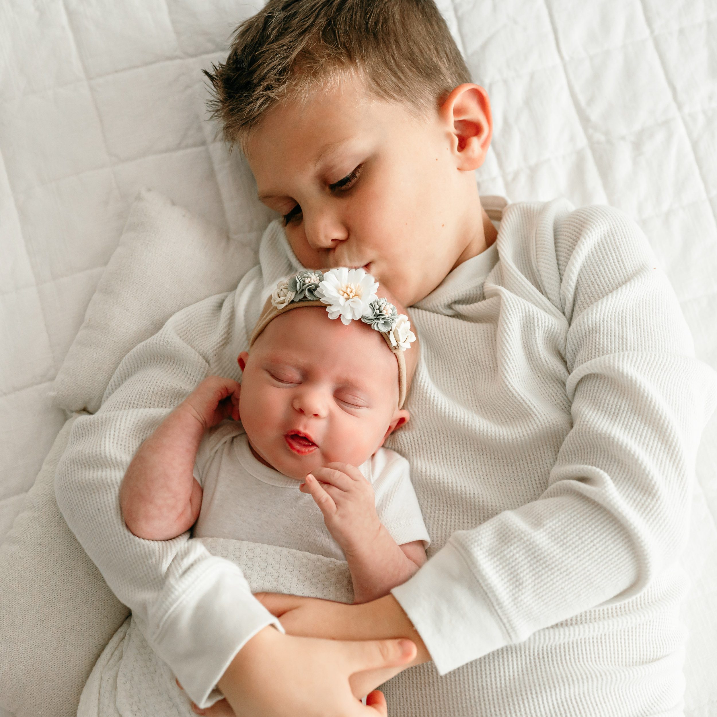 an older brother laying on a bed and kissing his baby sister on the head during a natural light newborn photo session
