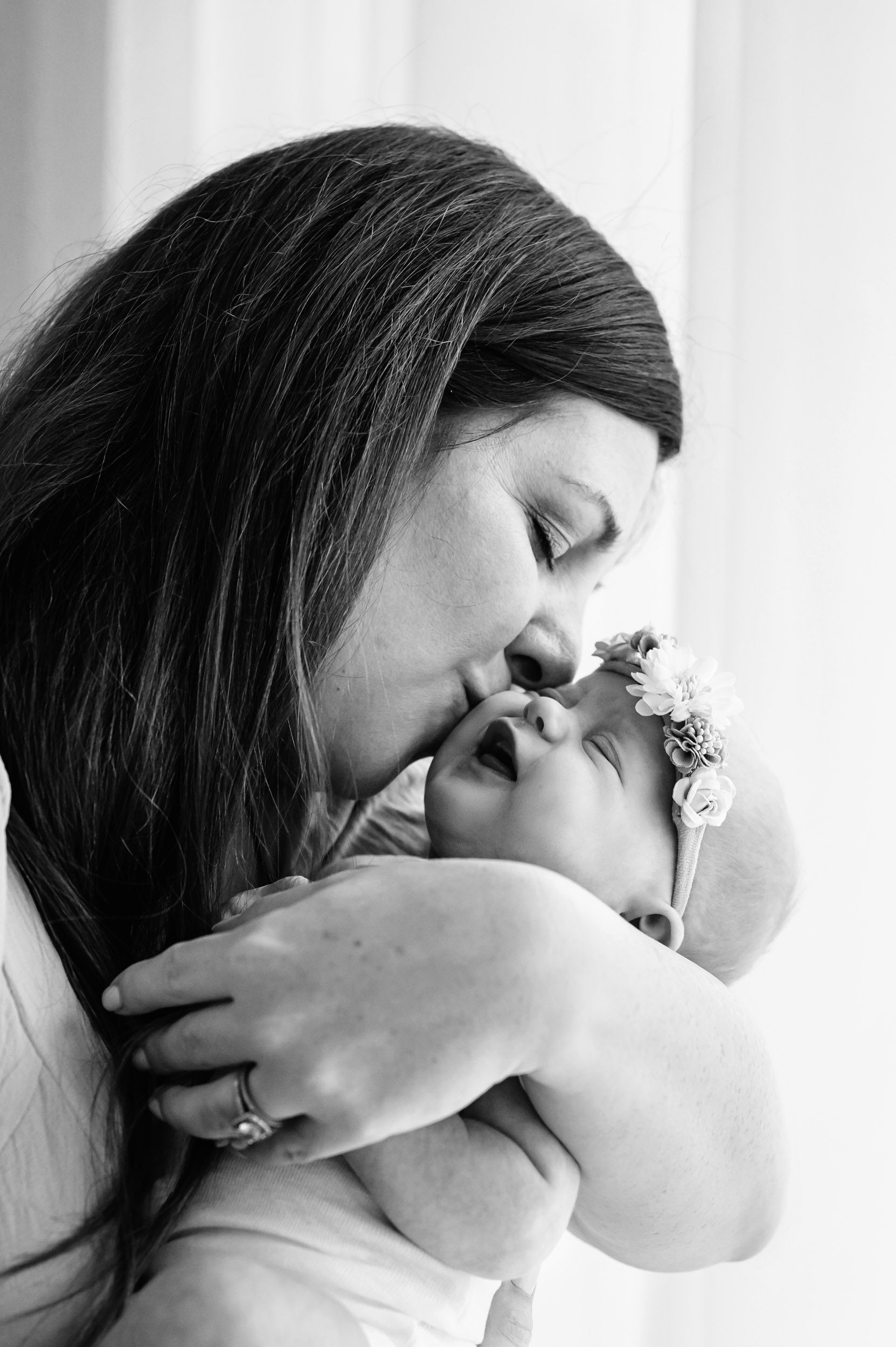 a black and white picture of a mom holding her baby girl and kissing her cheek while the baby smiles during a natural light newborn photoshoot