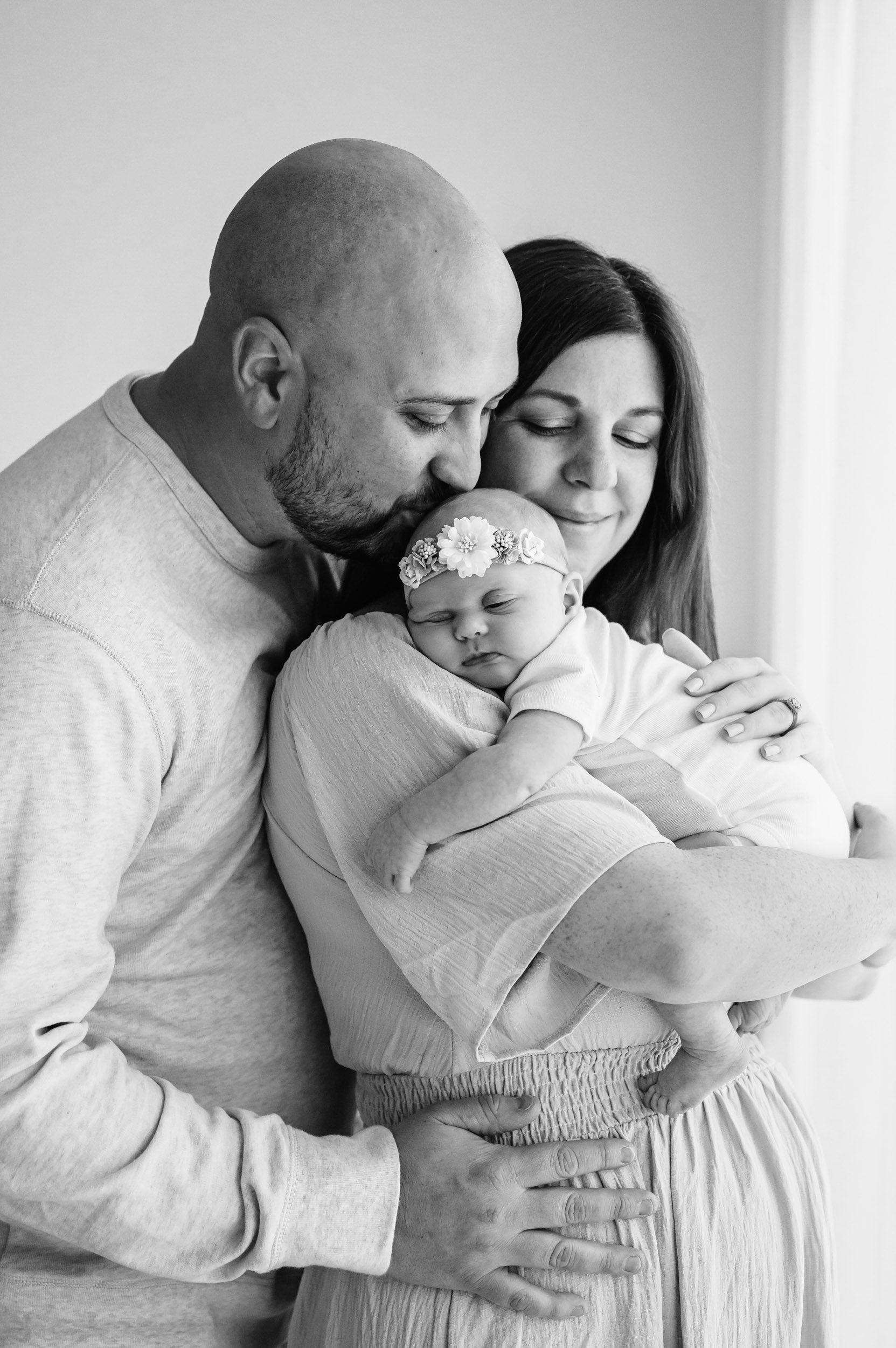 a black and white picture of a mom hugging her baby girl against her chest as dad hugs her from behind and gently kisses his daughter on the head during a newborn photo session
