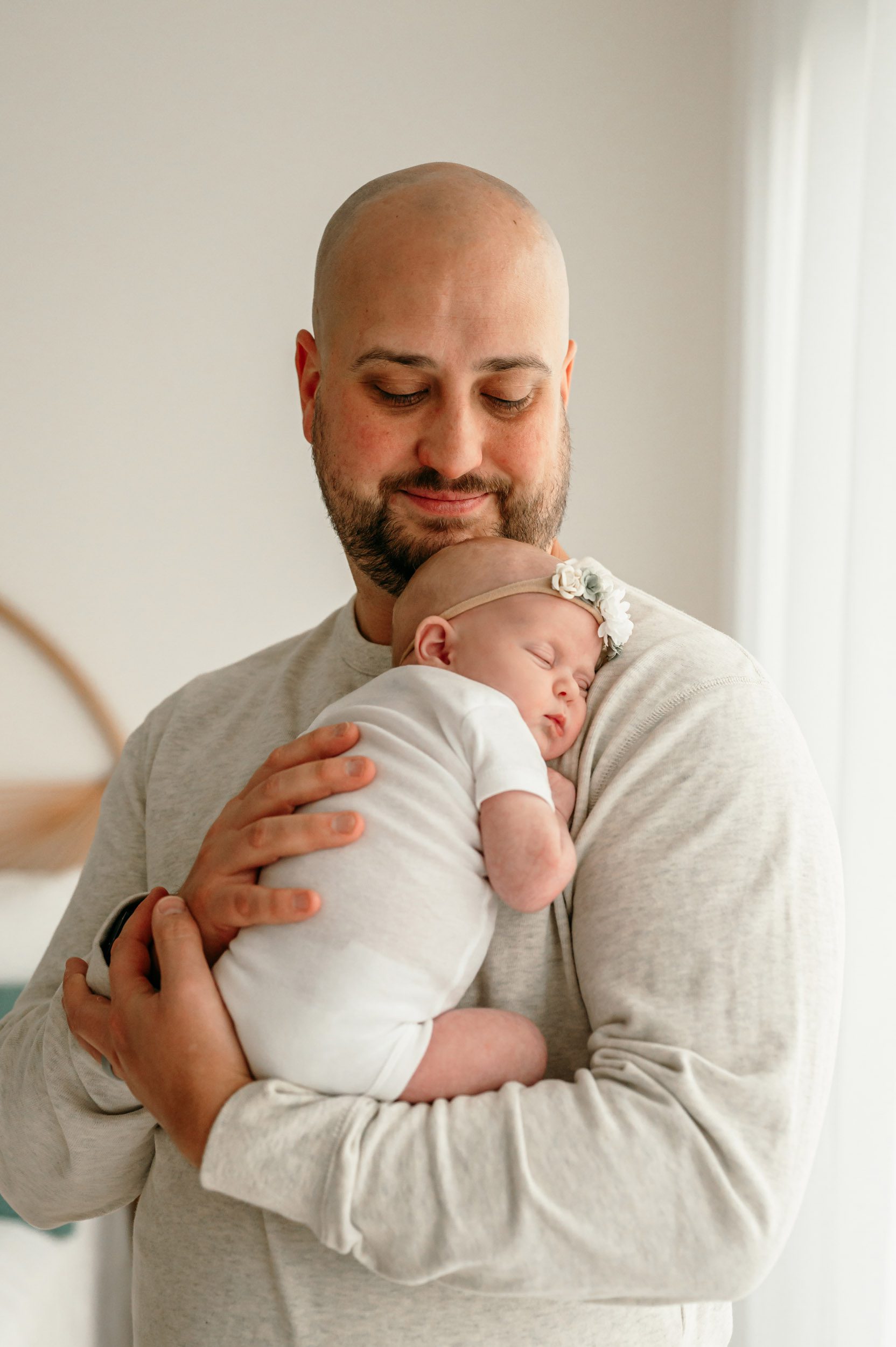 a father cradling his baby girl against his chest and smiling down at her during a natural light newborn photoshoot