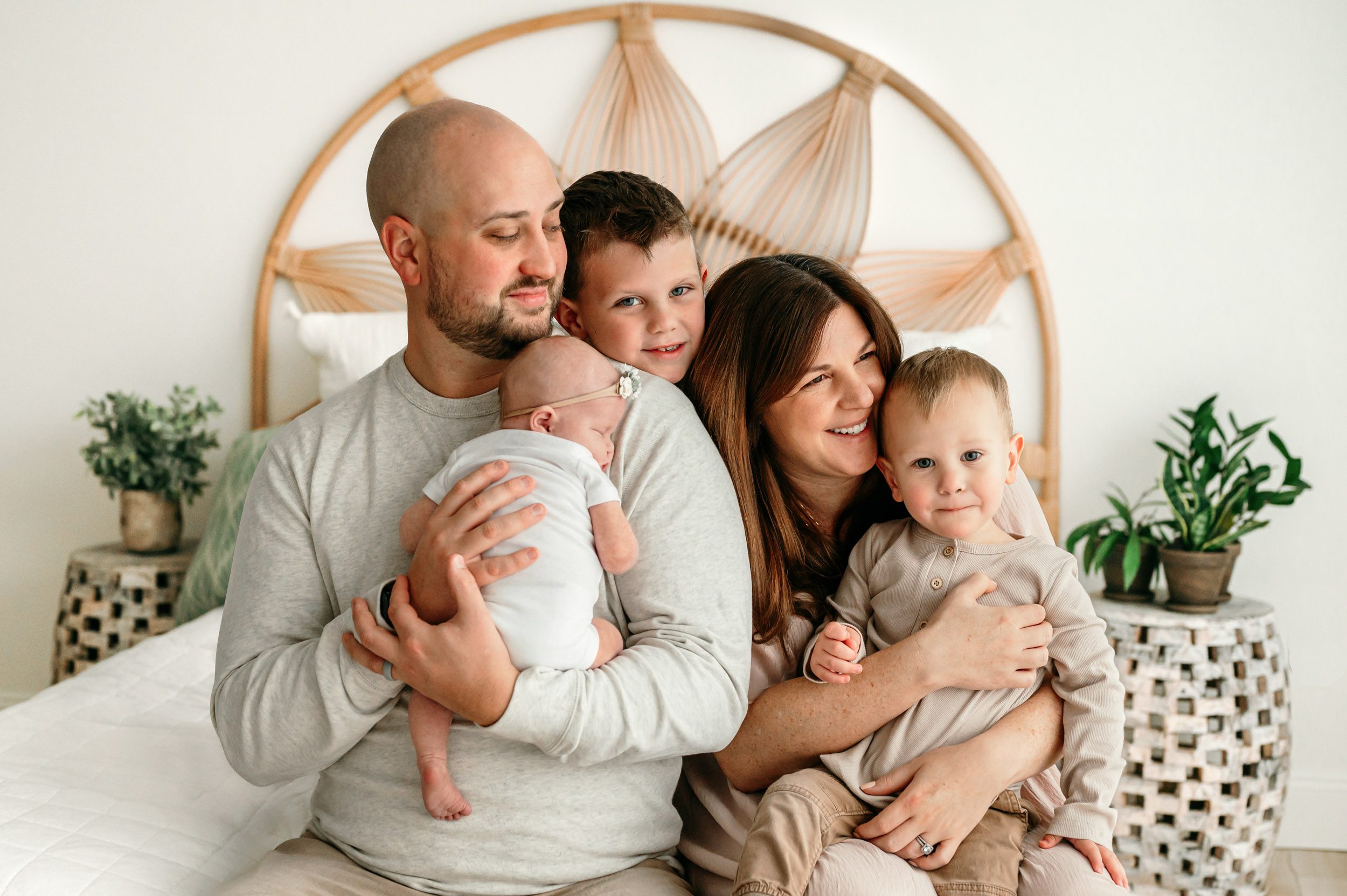 a mom and dad sitting on a bed while dad holds his baby girl in his arms and mom hugs one of her sons during a newborn photoshoot