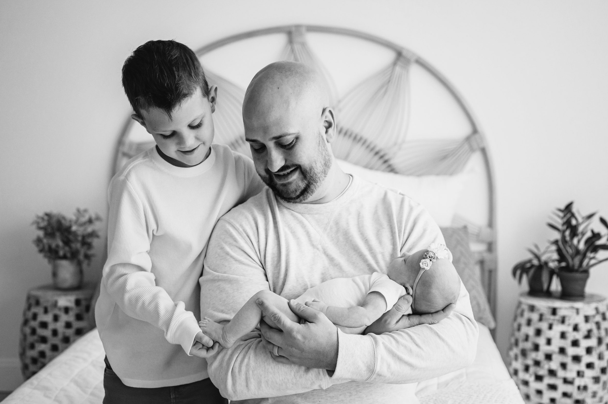 a black and white picture of a dad sitting on a bed and cradling his baby girl in his arms while his son hugs him and touches his baby sister's does during a natural light newborn photo session