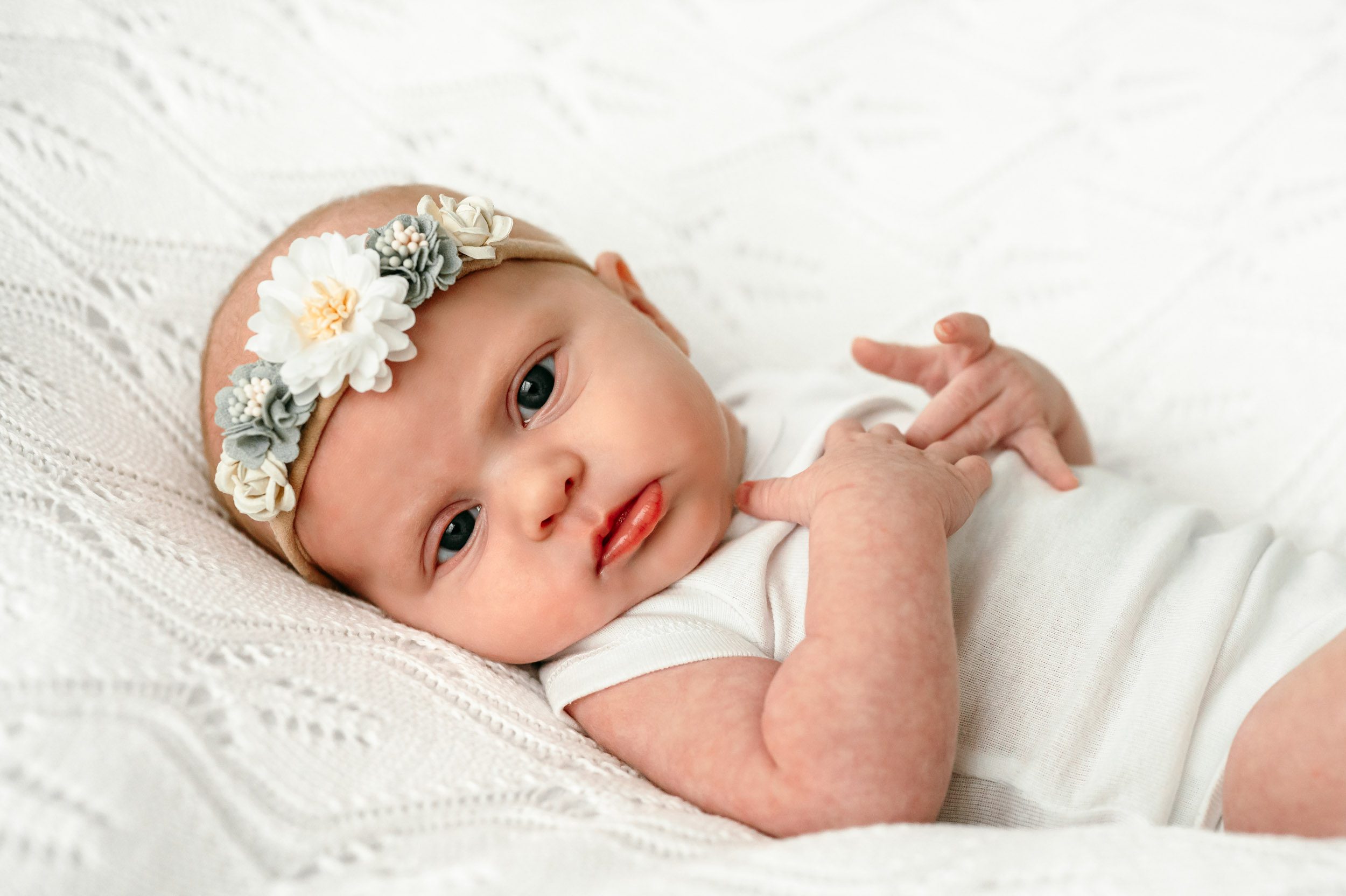 a baby girl in a white onesie and flower headband laying on a white textured backdrop and gazing at the camera with wide open eyes during a newborn photoshoot