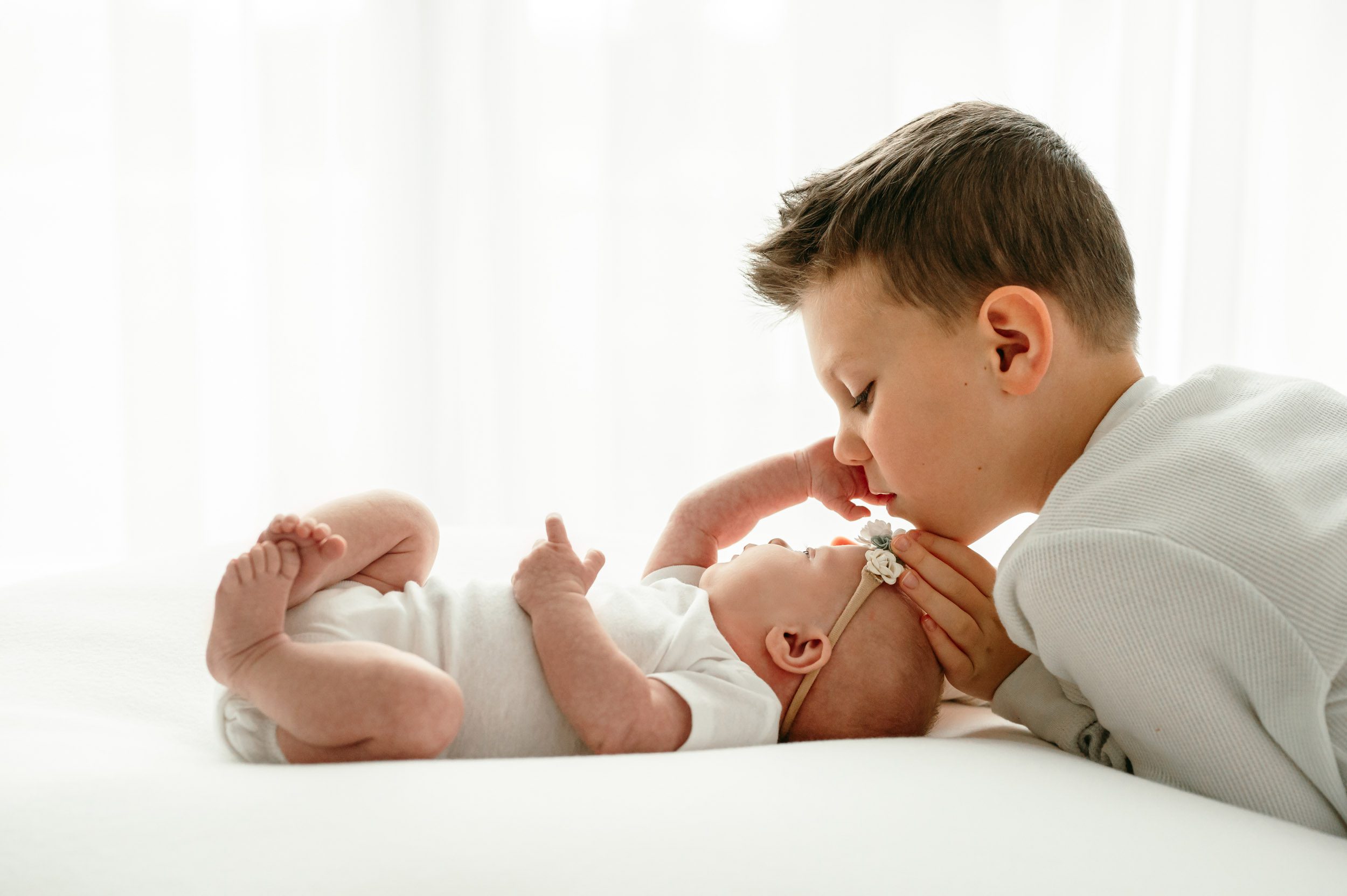 a backlit photo of a baby girl reaching up to touch her older brother's cheek as he gently touches her head during a natural light newborn photoshoot