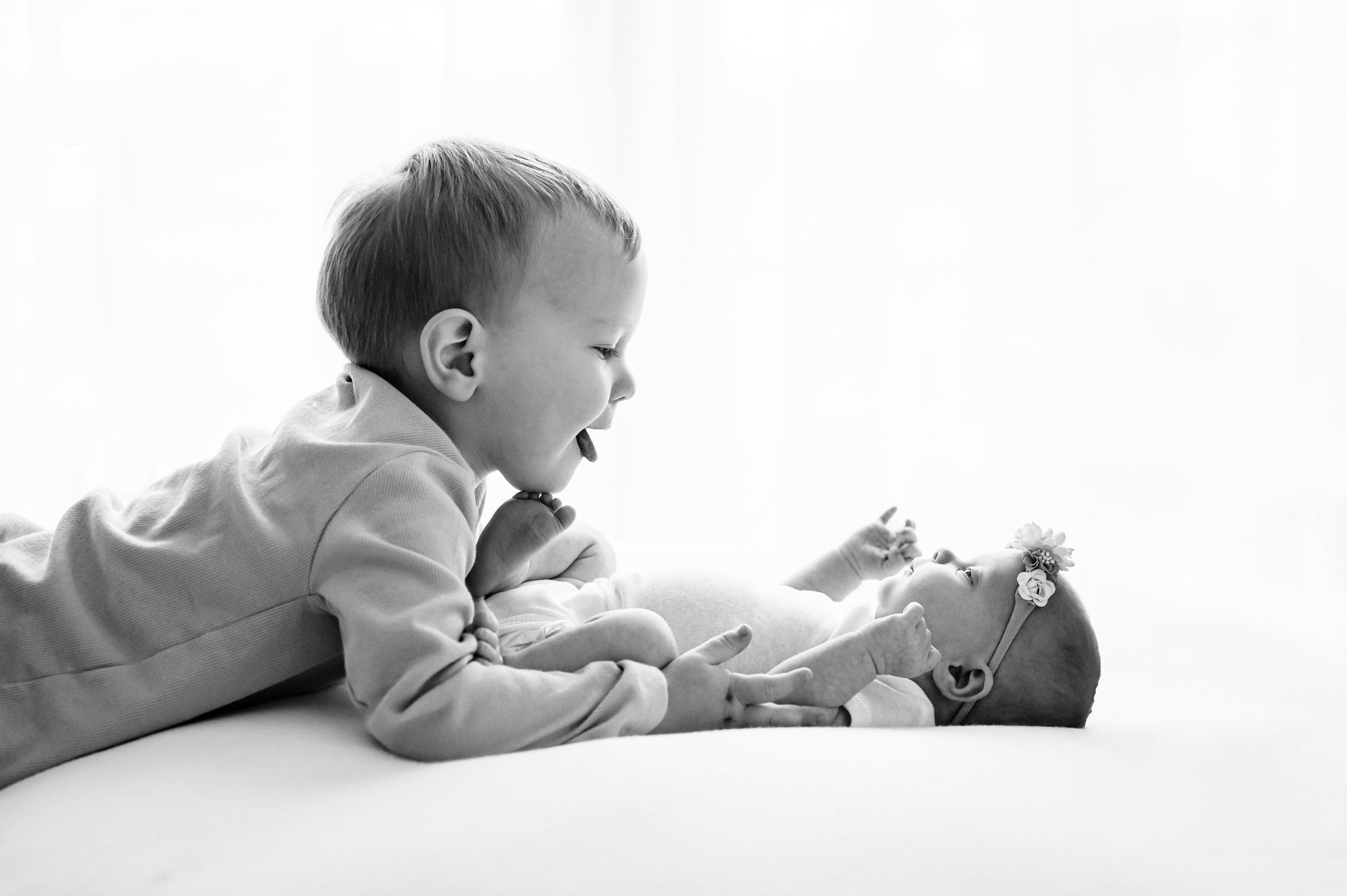 a black and white backlit photo of a baby girl laying on a white backdrop as her older brother hugs her and sticks his tongue out at her during a natural light newborn photoshoot