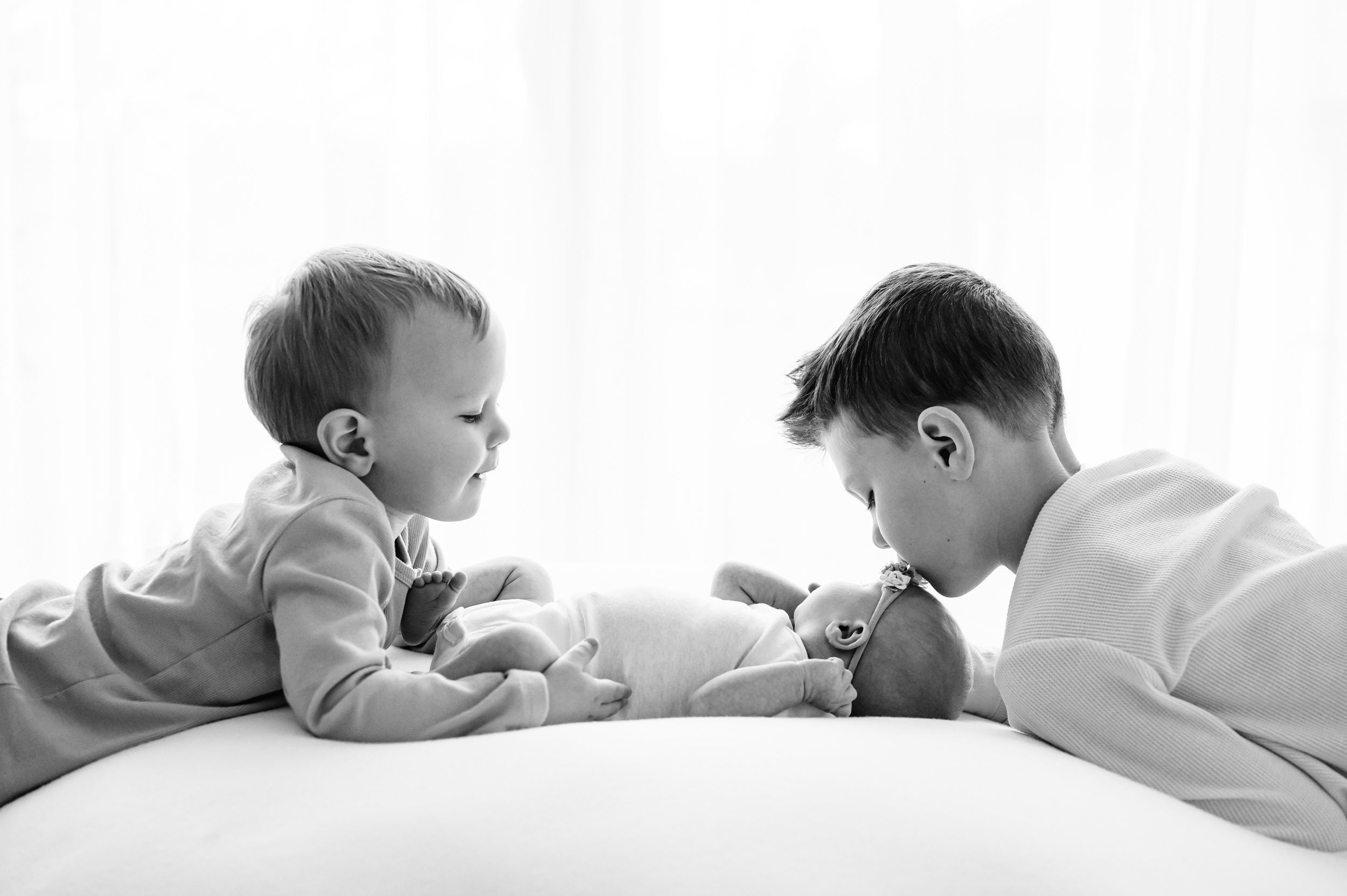 a black and white backlit photo of a baby girl laying on a white backdrop with her two older brothers kissing and hugging her during a natural light newborn photo session