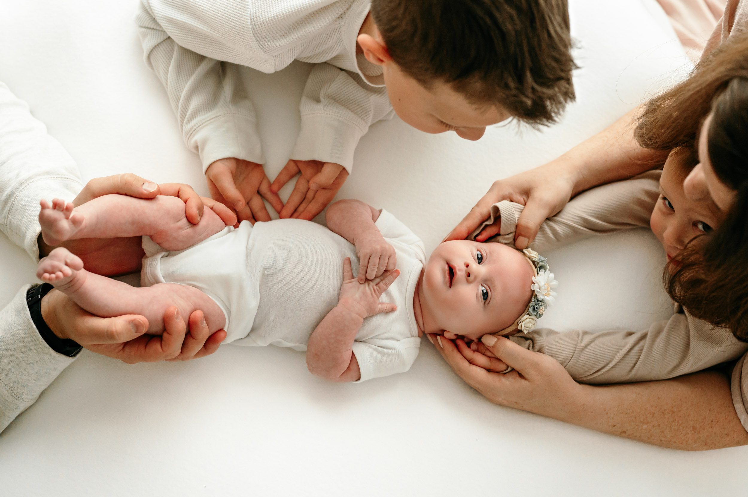 a baby girl laying on a white backdrop and looking up at the camera while her parents and two big brothers touch her with their hands during a natural light newborn photoshoot