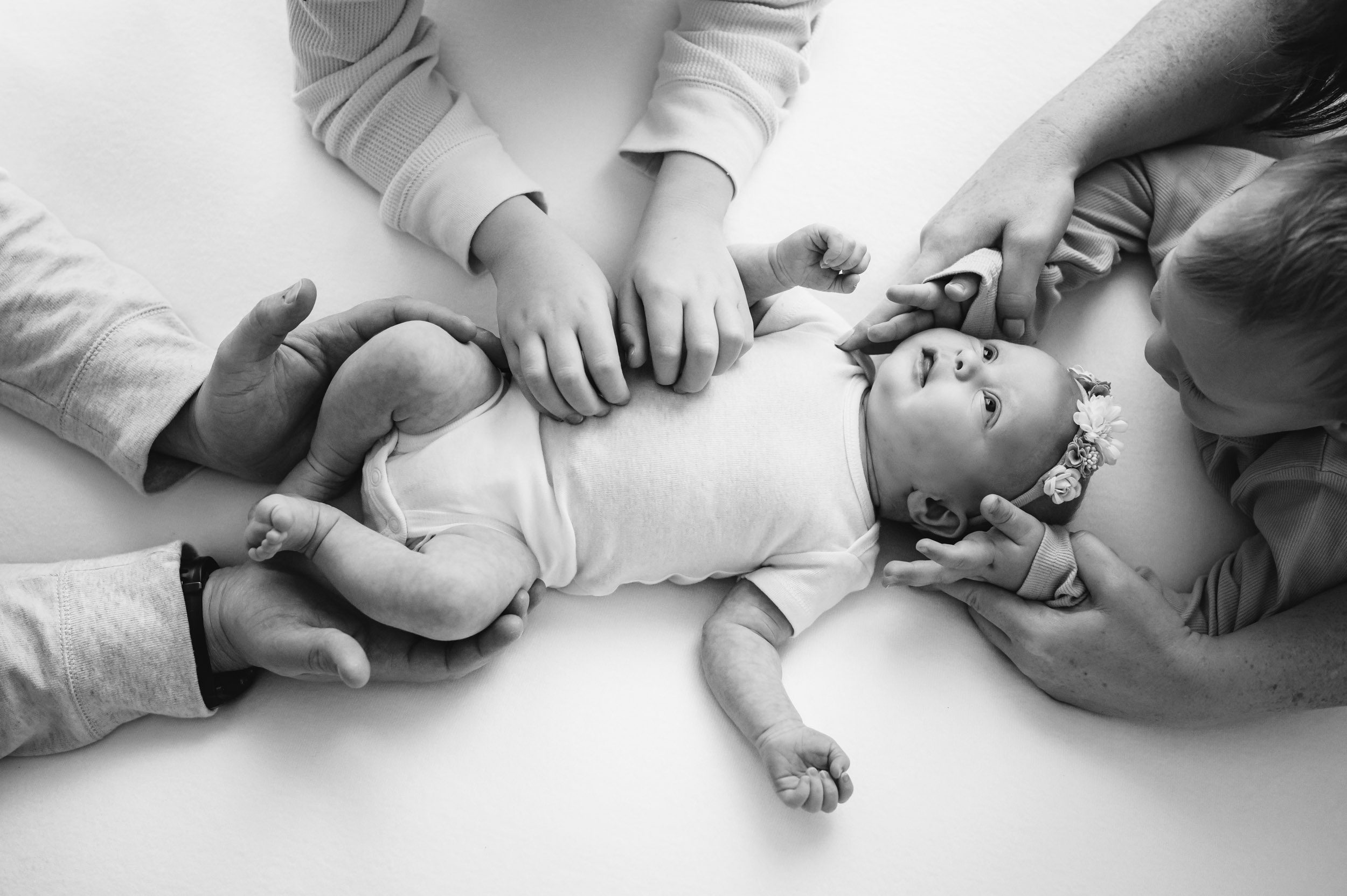 a black and white picture of a baby girl laying on a white backdrop with her parents and two older brothers all around her touching her with their hands during a newborn photoshoot