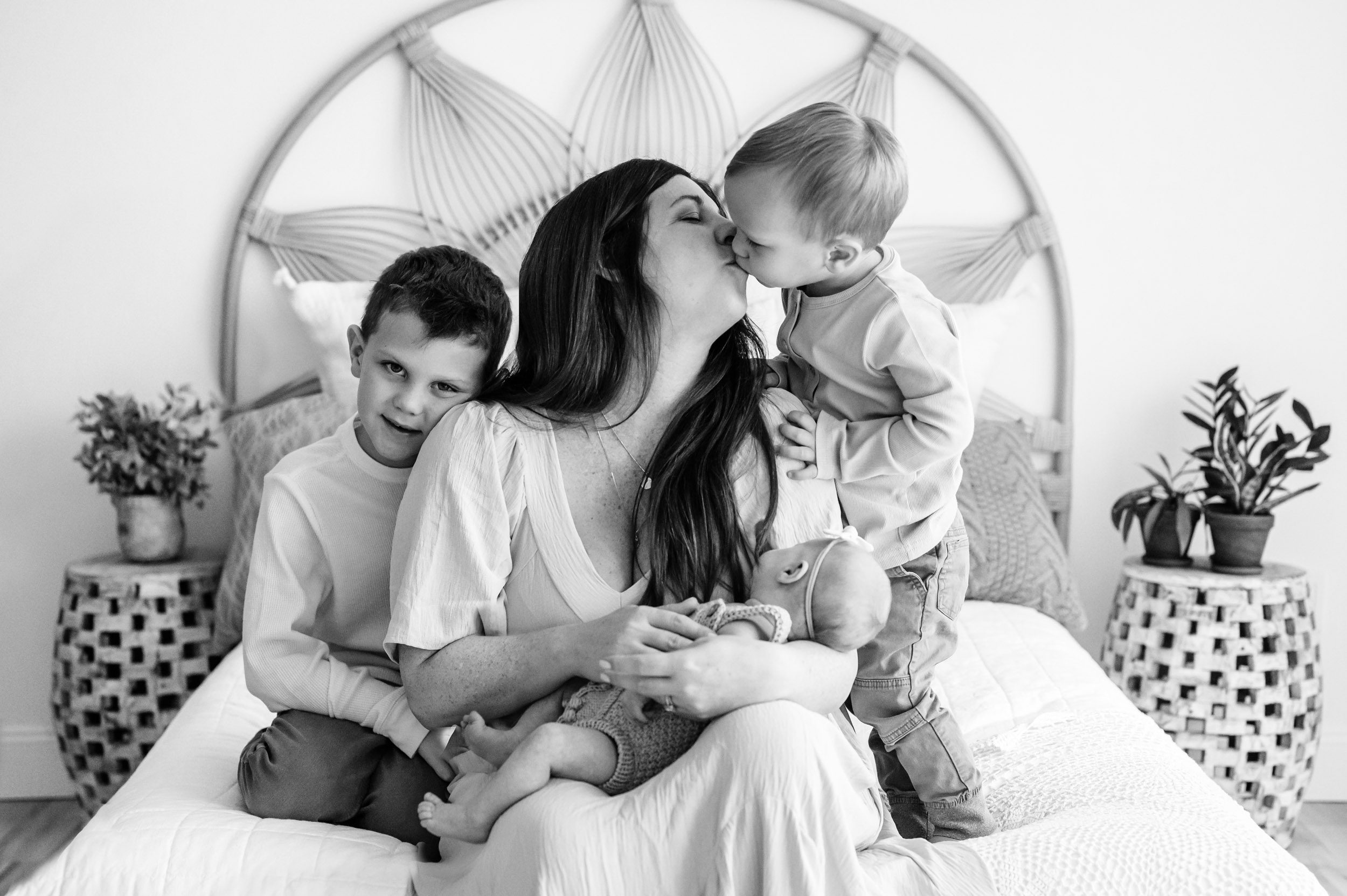 a black and white picture of a mom holding her baby girl in her arms and kissing her youngest son as her older son hugs her arm during a natural light newborn photo session