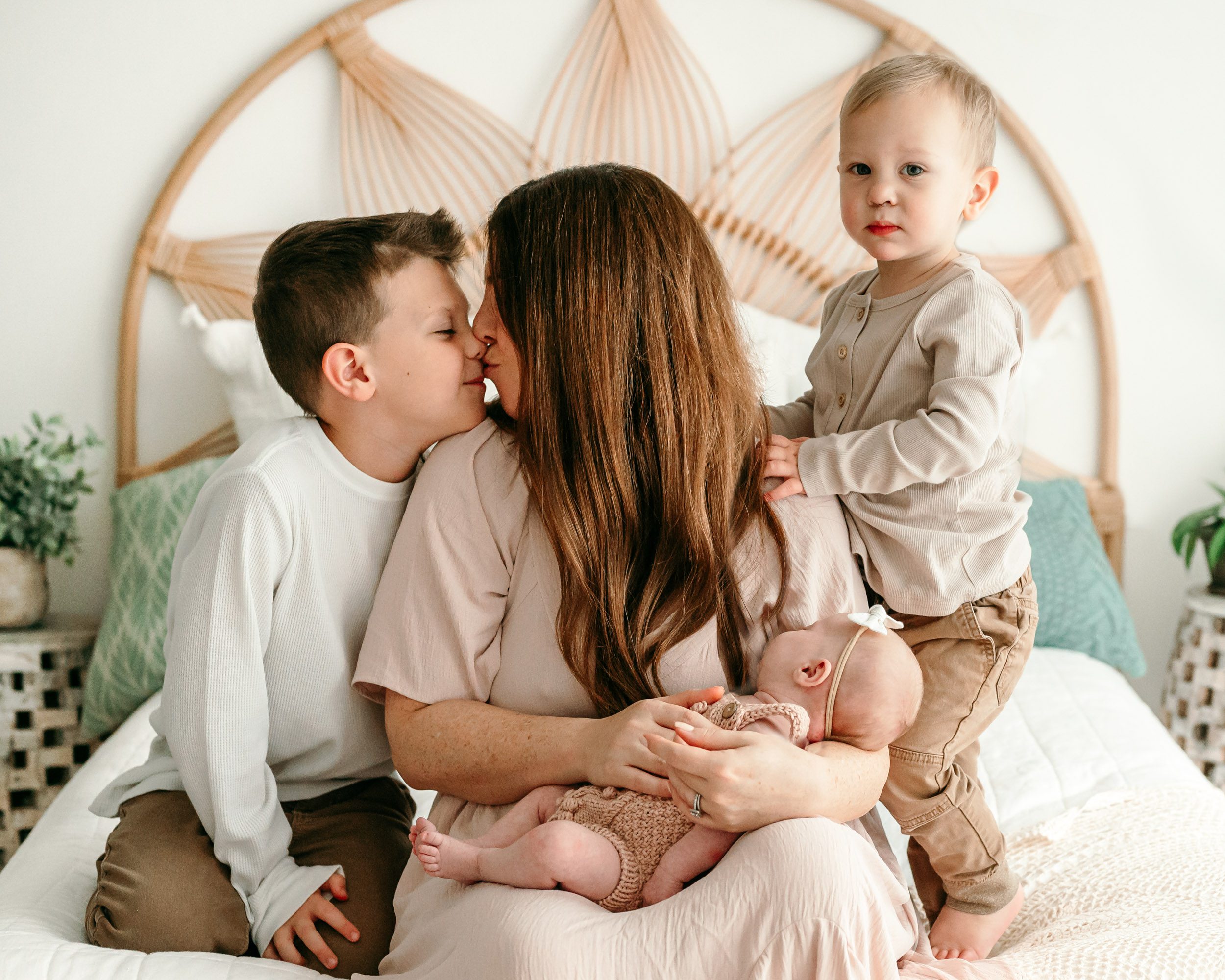 a mom sitting on a bed with her three kids and kissing her oldest son on the nose while she holds her baby girl in her arms and her younger son hugs her arm during a Pottstown newborn photoshoot