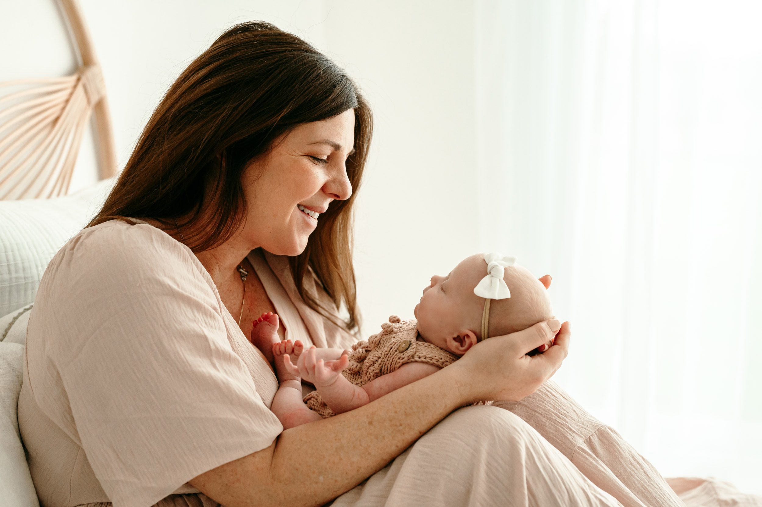 a new mom sitting on a bed holding her baby girl and smiling down at her during a newborn photoshoot