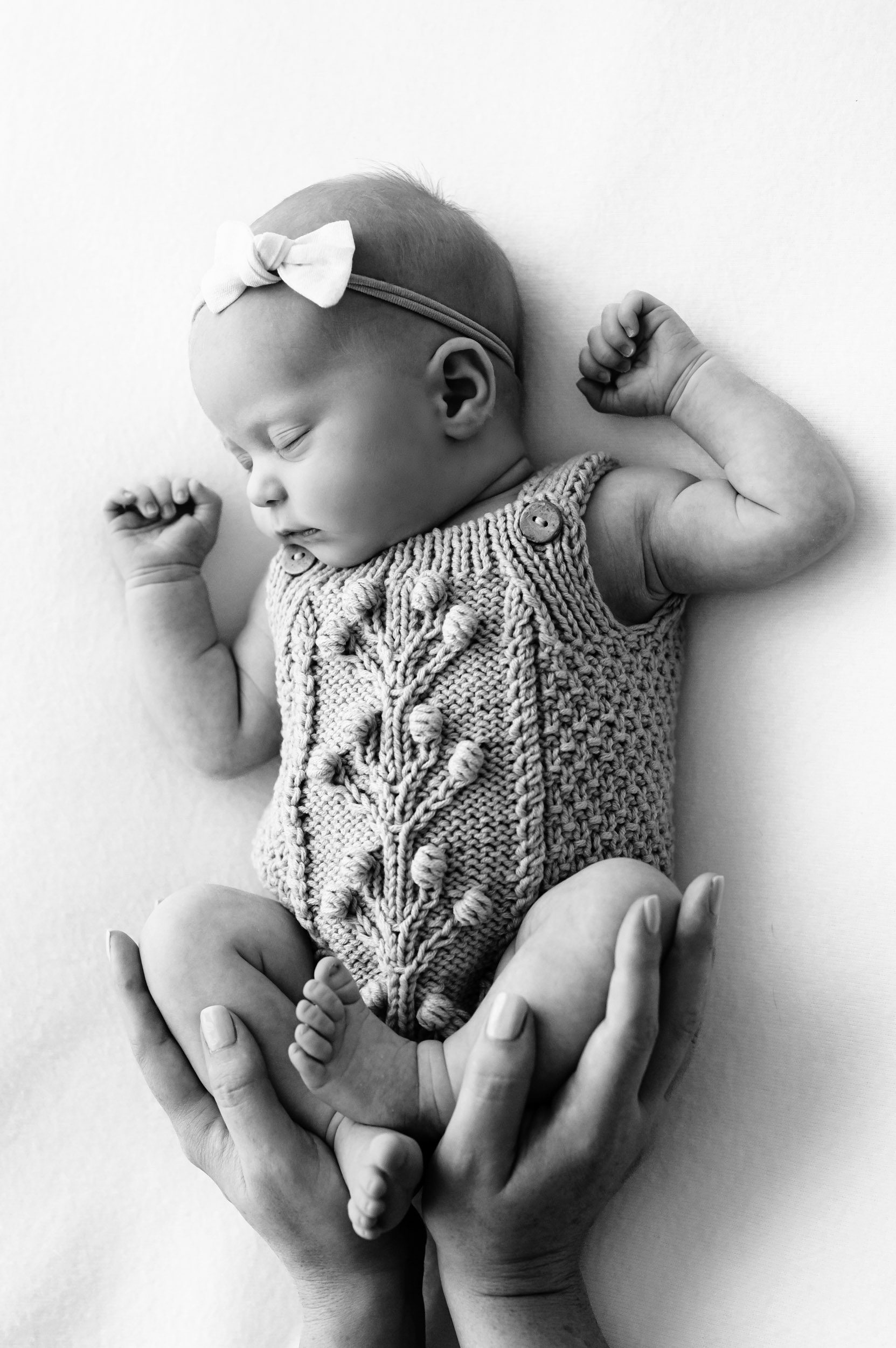 a black and white photo of a baby girl sleeping on a white backdrop while her mom cradles her legs in her hands during a newborn photo session