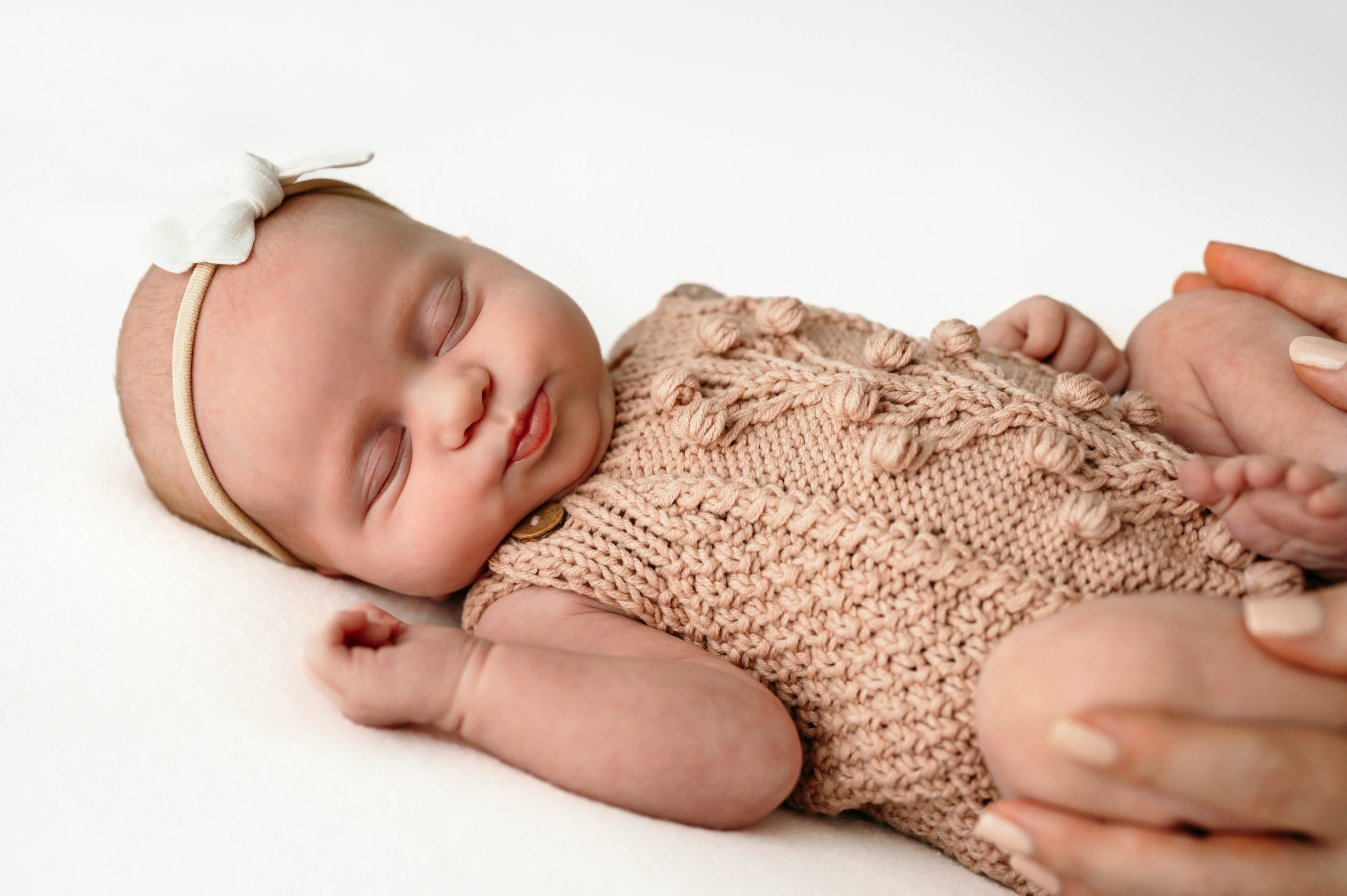 a baby girl in a pink knit romper and white headband sleeping on a white backdrop and looking toward the camera with pursed lips while her mom holds her legs in her hands during a newborn photoshoot