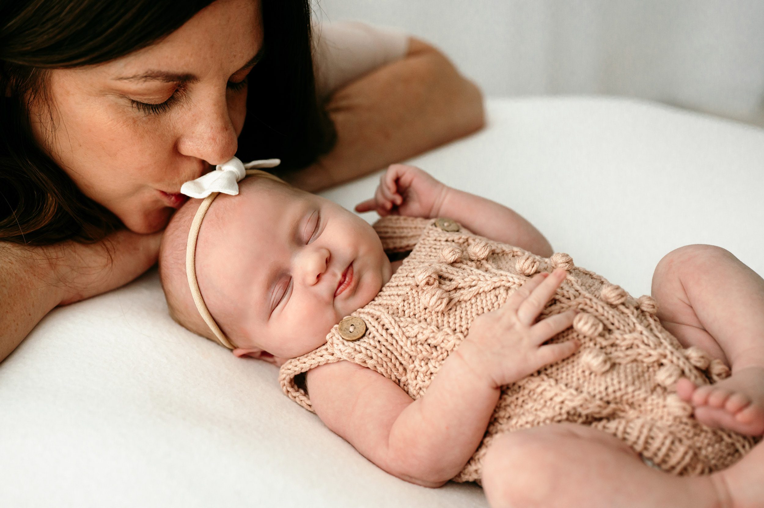 a baby girl sleeping on a white blanket as her mom gently kisses her on the head during a newborn photoshoot