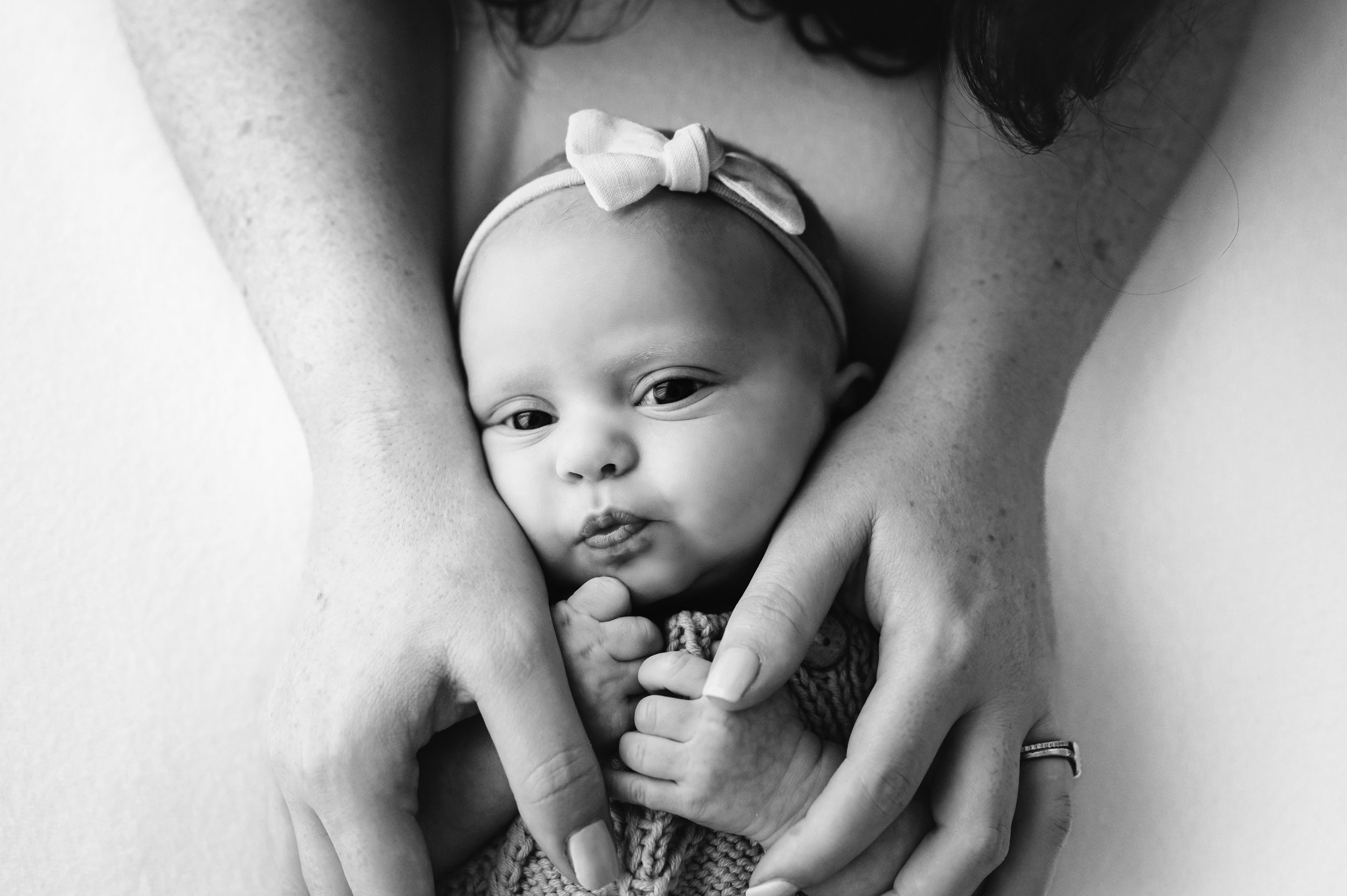 a black and white picture of a baby girl laying on a white backdrop and gazing up at the camera with her mom's hands cradling her face during a newborn photo session
