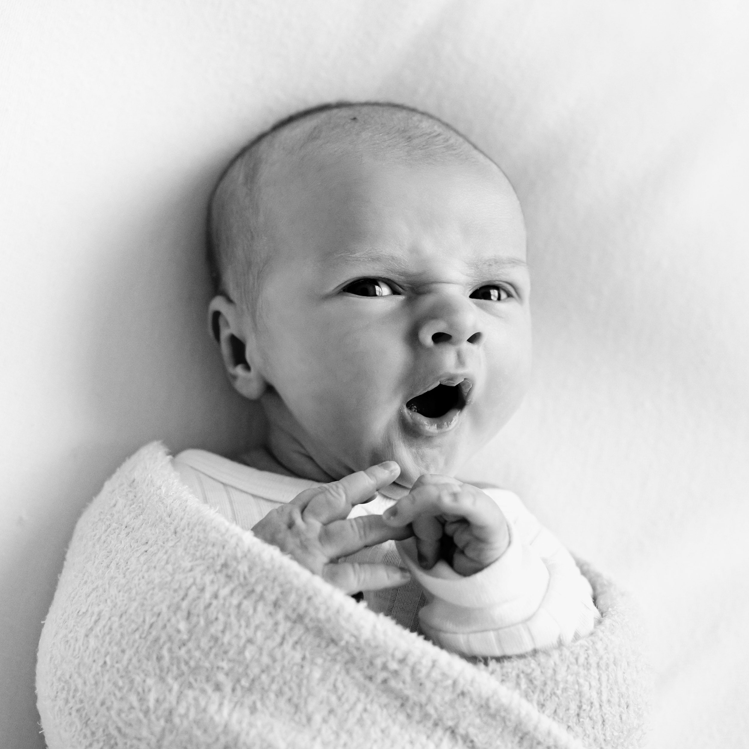 a black and white picture of a newborn baby boy looking at the camera and yawning as he touches his hands together during a newborn photoshoot