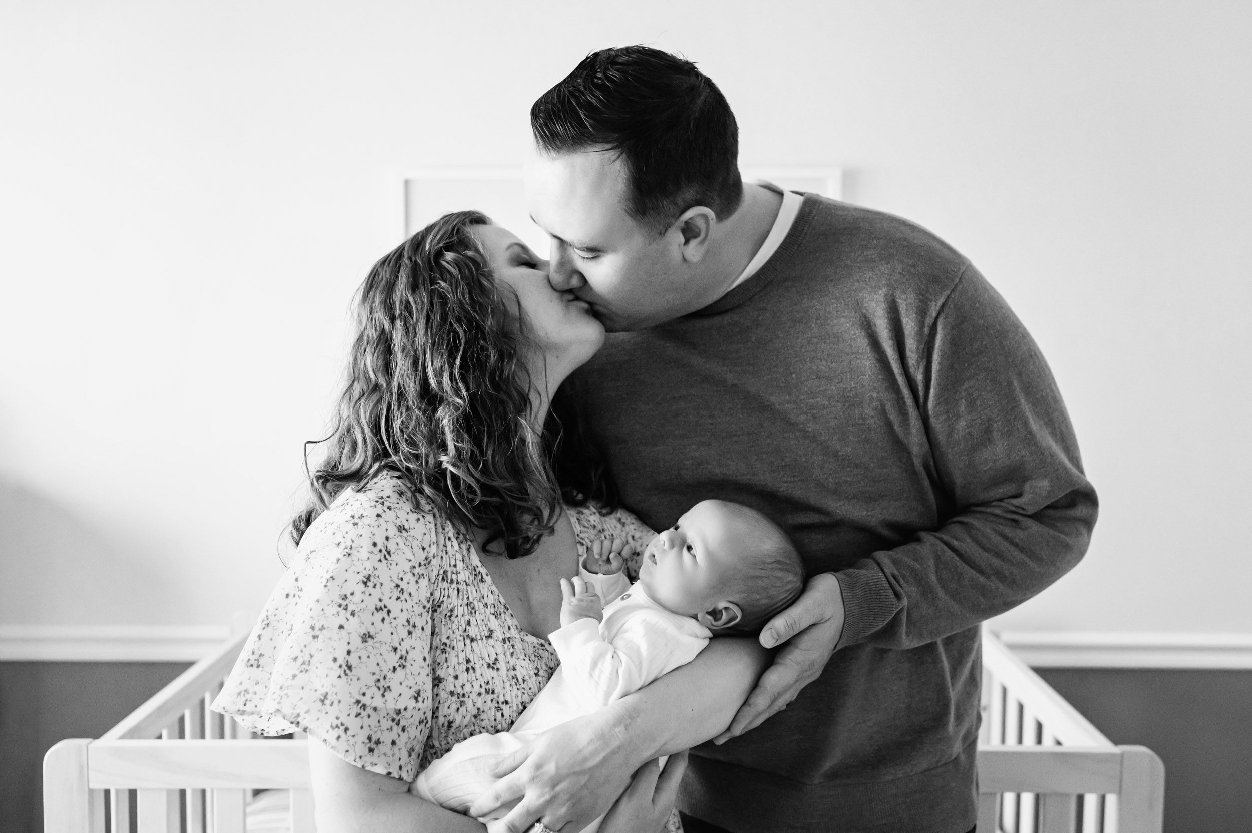 a black and white picture of a mom and dad standing in front of the crib in the nursery holding their newborn son in their arms and kissing each other during a newborn baby photography session