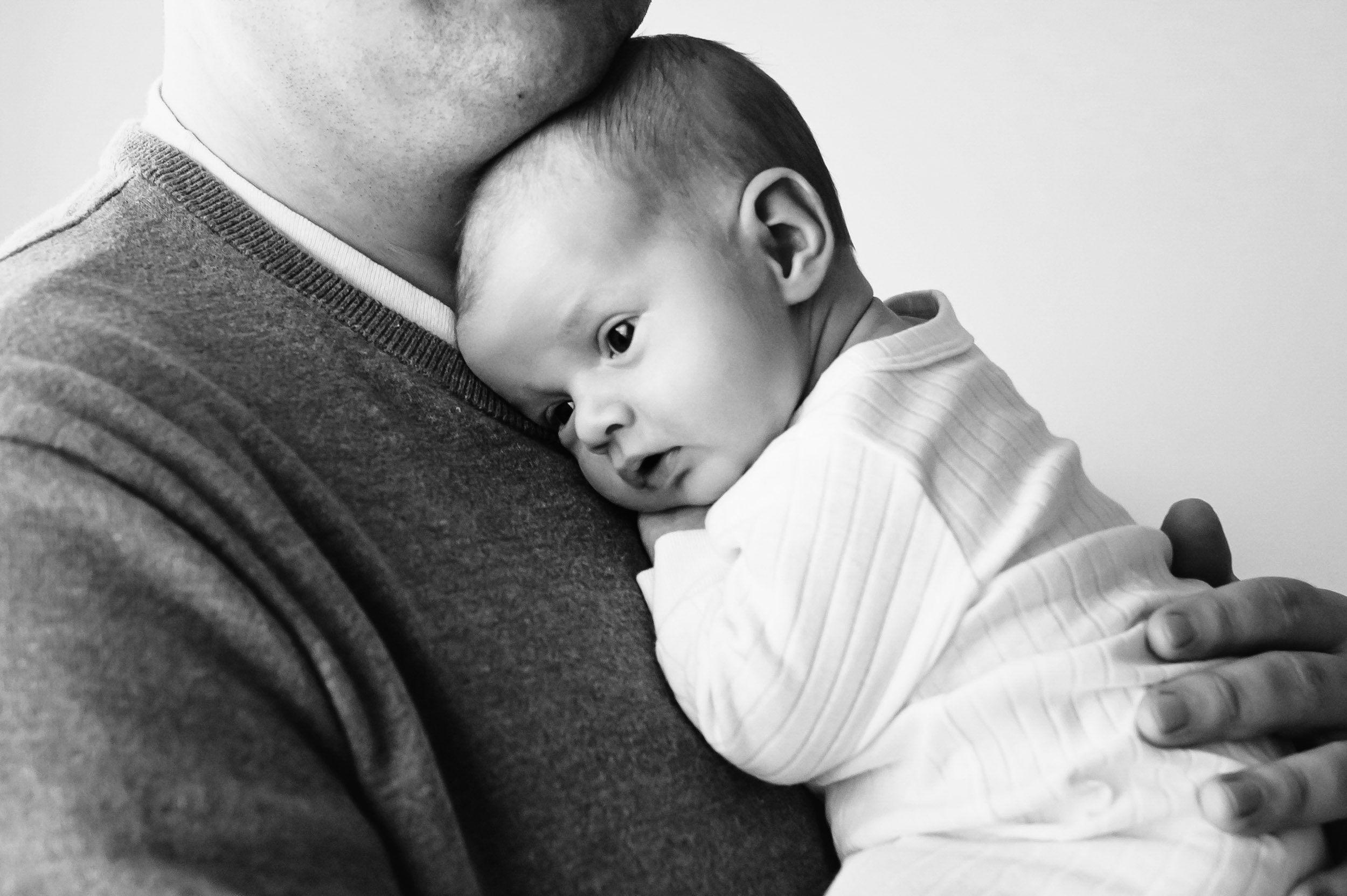 a black and white close up picture of newborn baby boy resting his head against his father's chest and looking toward the light coming in through the window during a newborn baby photography session