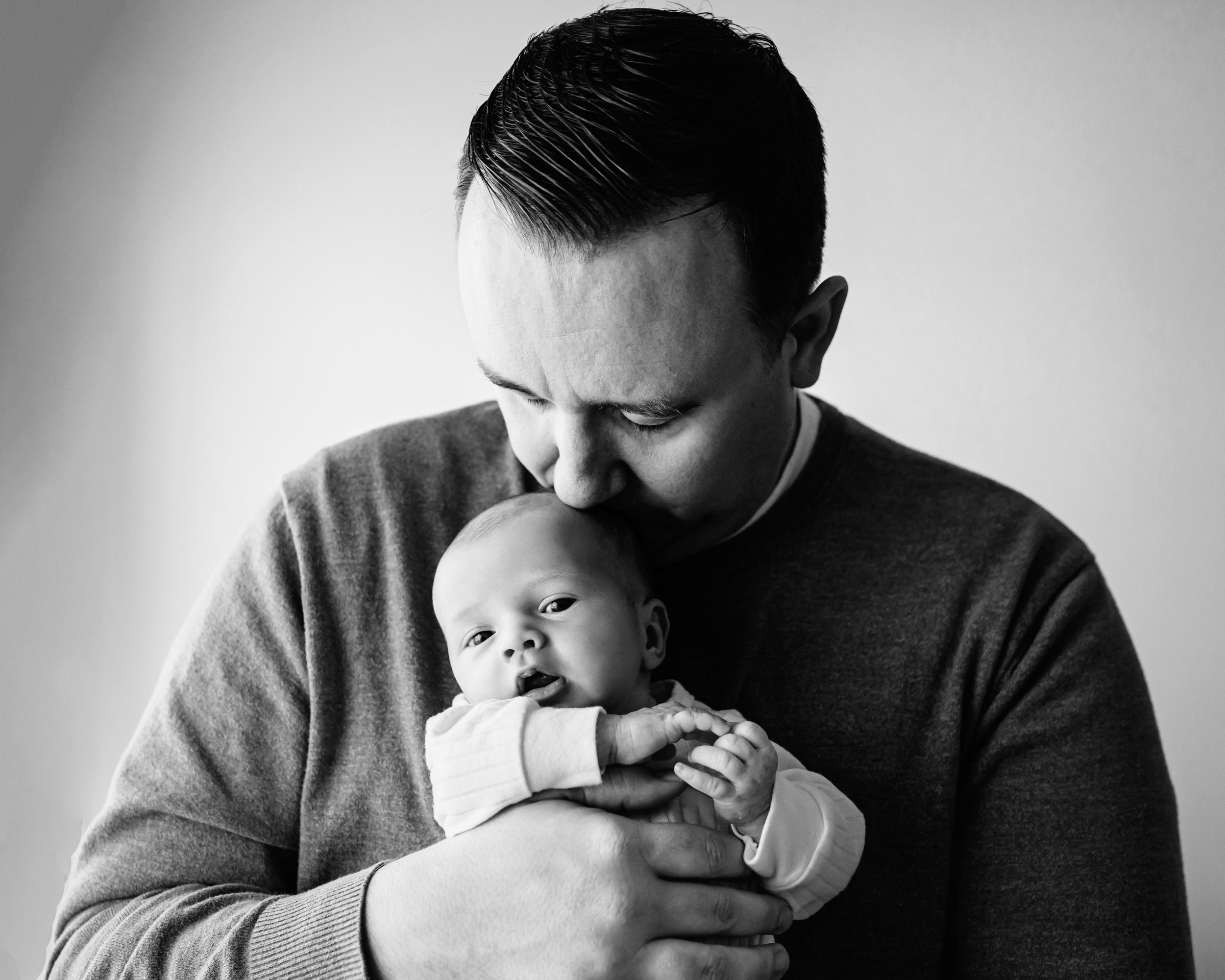 a black and white picture of dad holding his baby boy against his chest and kissing him on the head during a newborn baby photoshoot