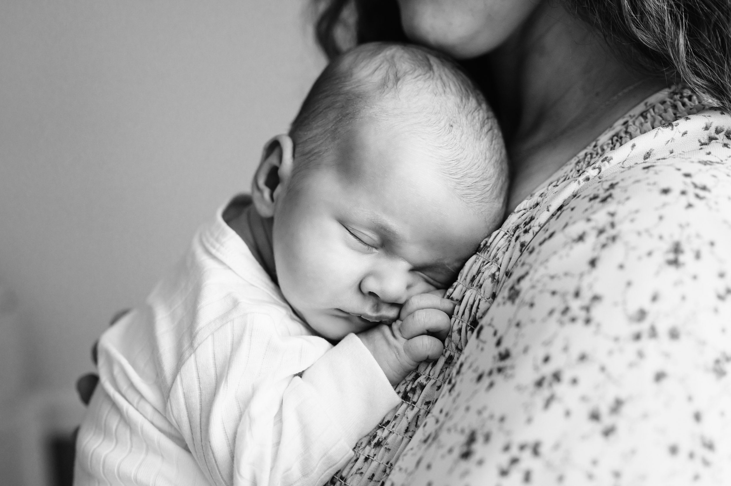 a black and white close up picture of baby boy wearing a long sleeve white onesie sleeping with his head resting  against his mom's chest during a newborn photos session