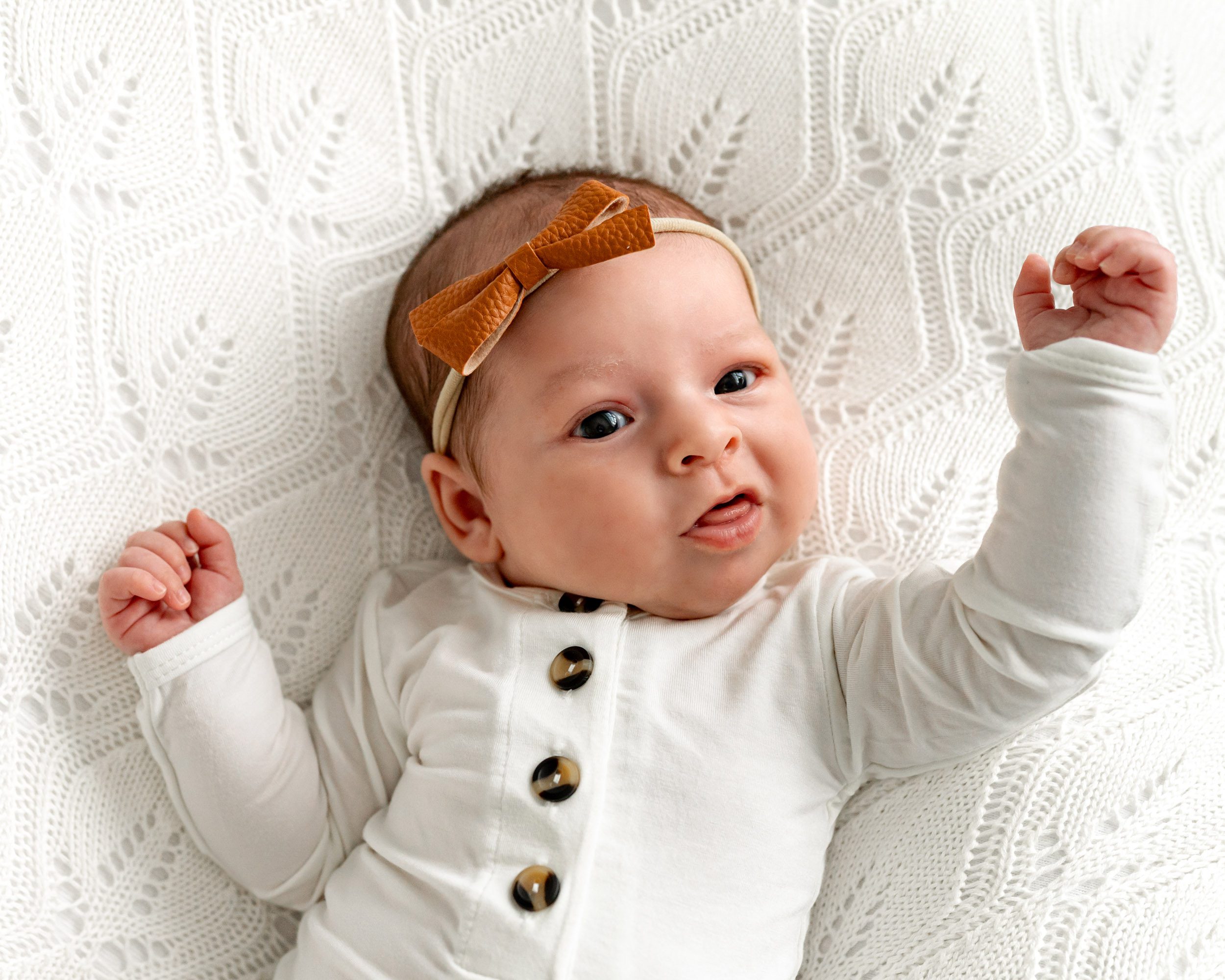 a baby girl in a white body suit laying on a white textured backdrop and lifting her arms up as she sticks out her tongue during a newborn photography session