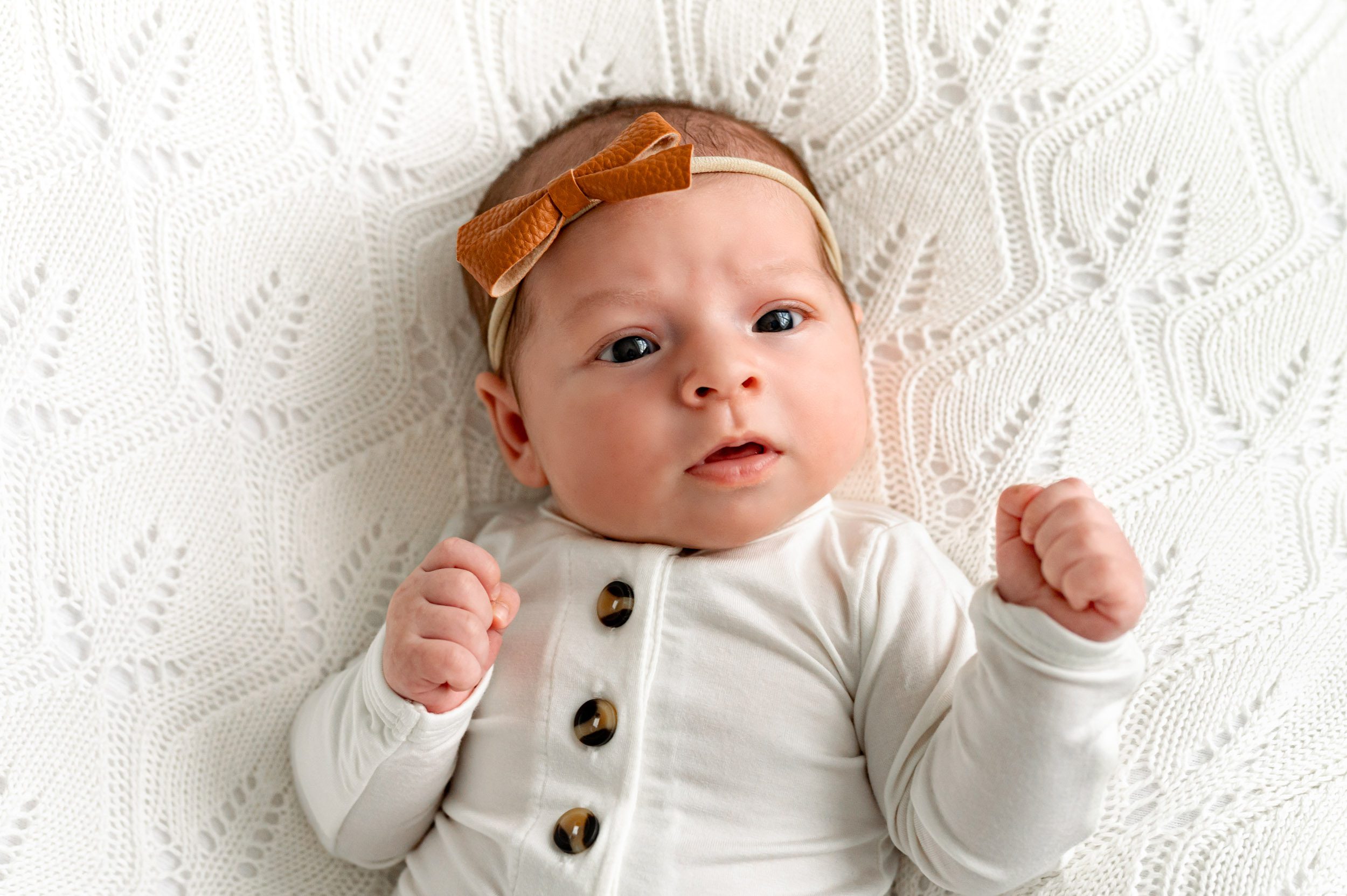 a baby girl in a white body suit laying on a white backdrop looking up toward the camera with wide open eyes during a newborn photoshoot