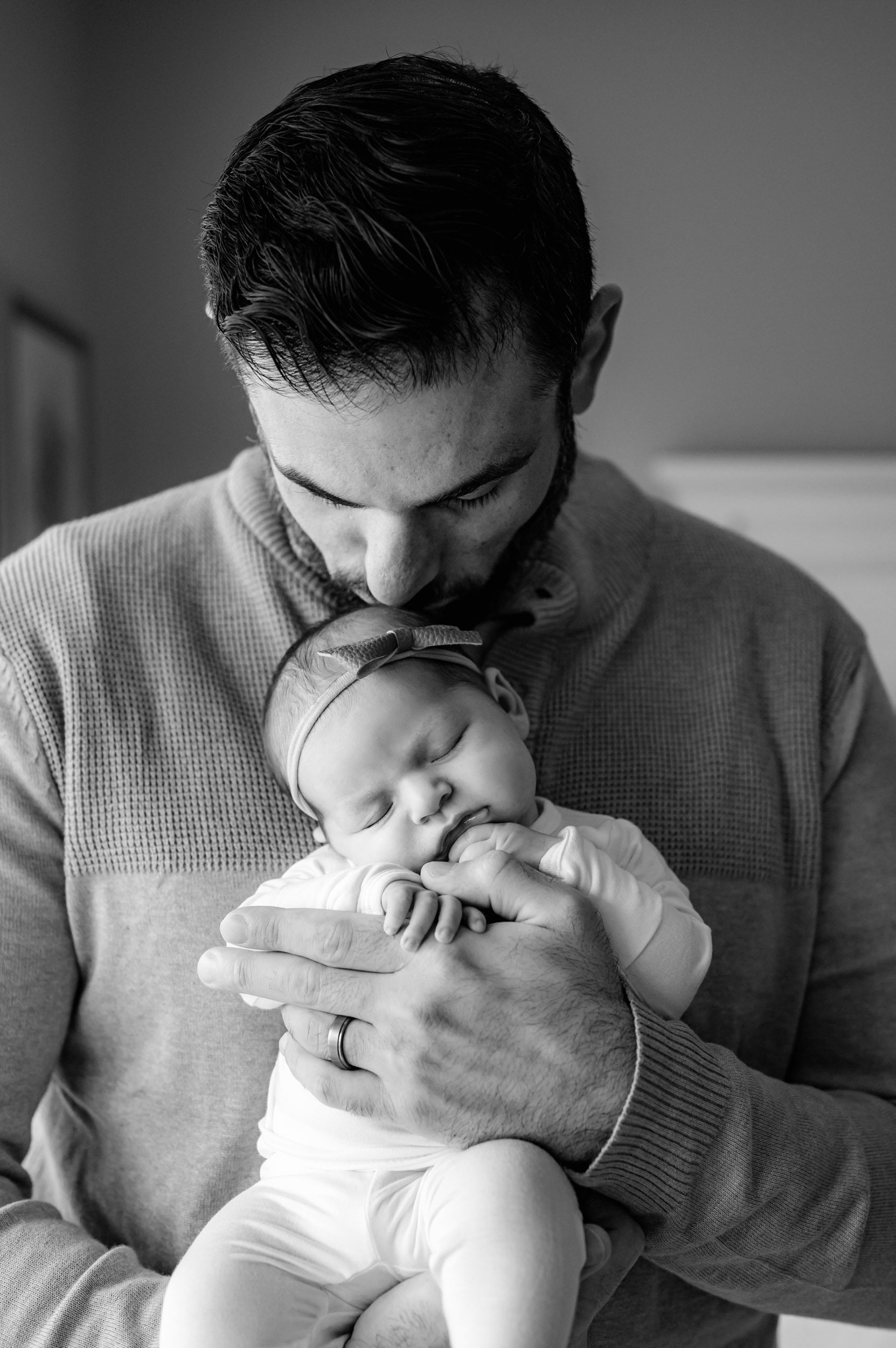 a black and white picture of a dad holding his sleeping baby girl in his hands as she grabs his hand with her fingers during a newborn photoshoot
