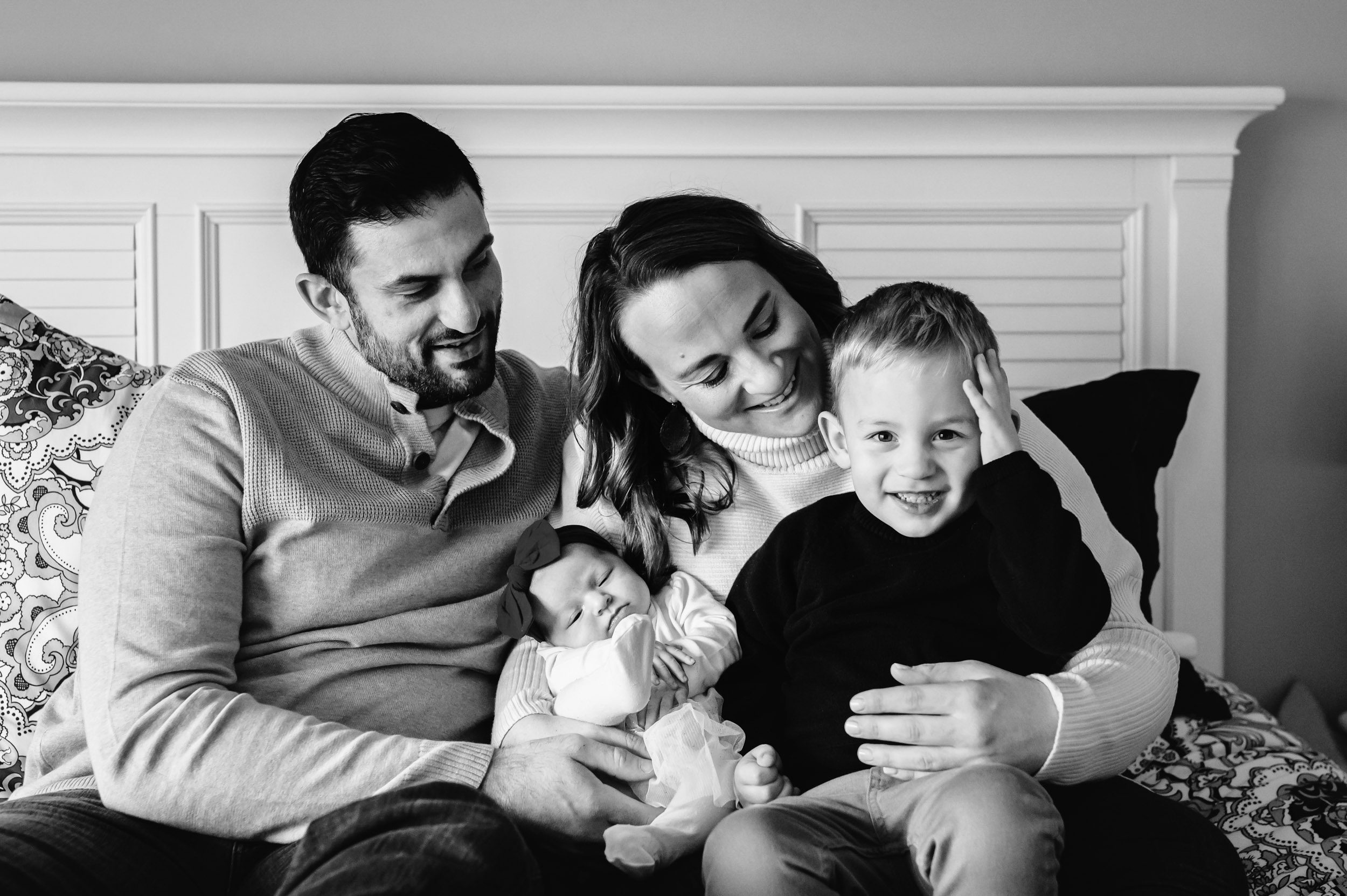 a mom and dad snuggling on a bed with their baby girl and young son in their arms as big brother smiles and holds his hand against his head during a newborn photography session