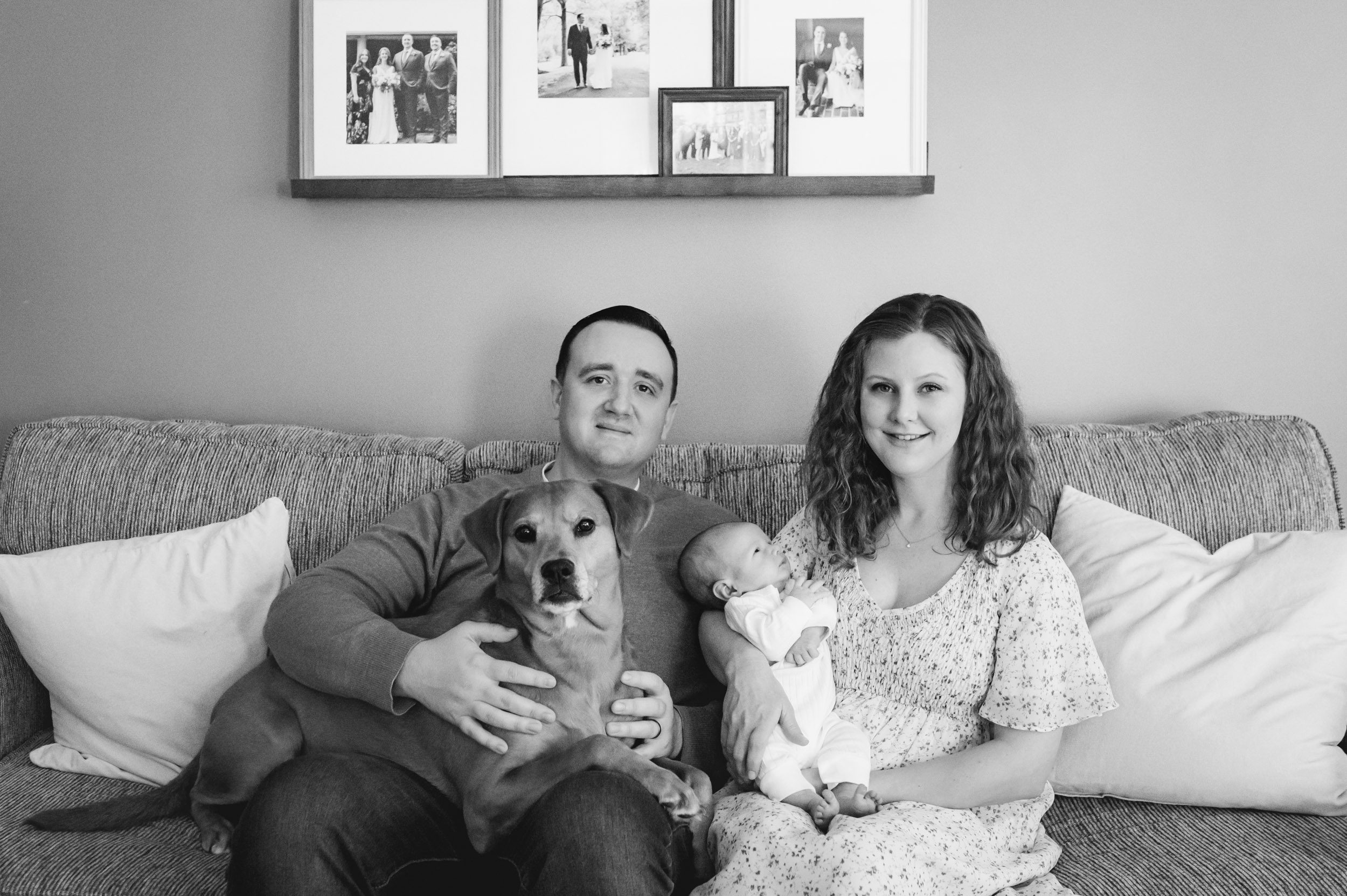 a black and white picture of new parents sitting on their couch with their baby boy and pet dog on their lap 