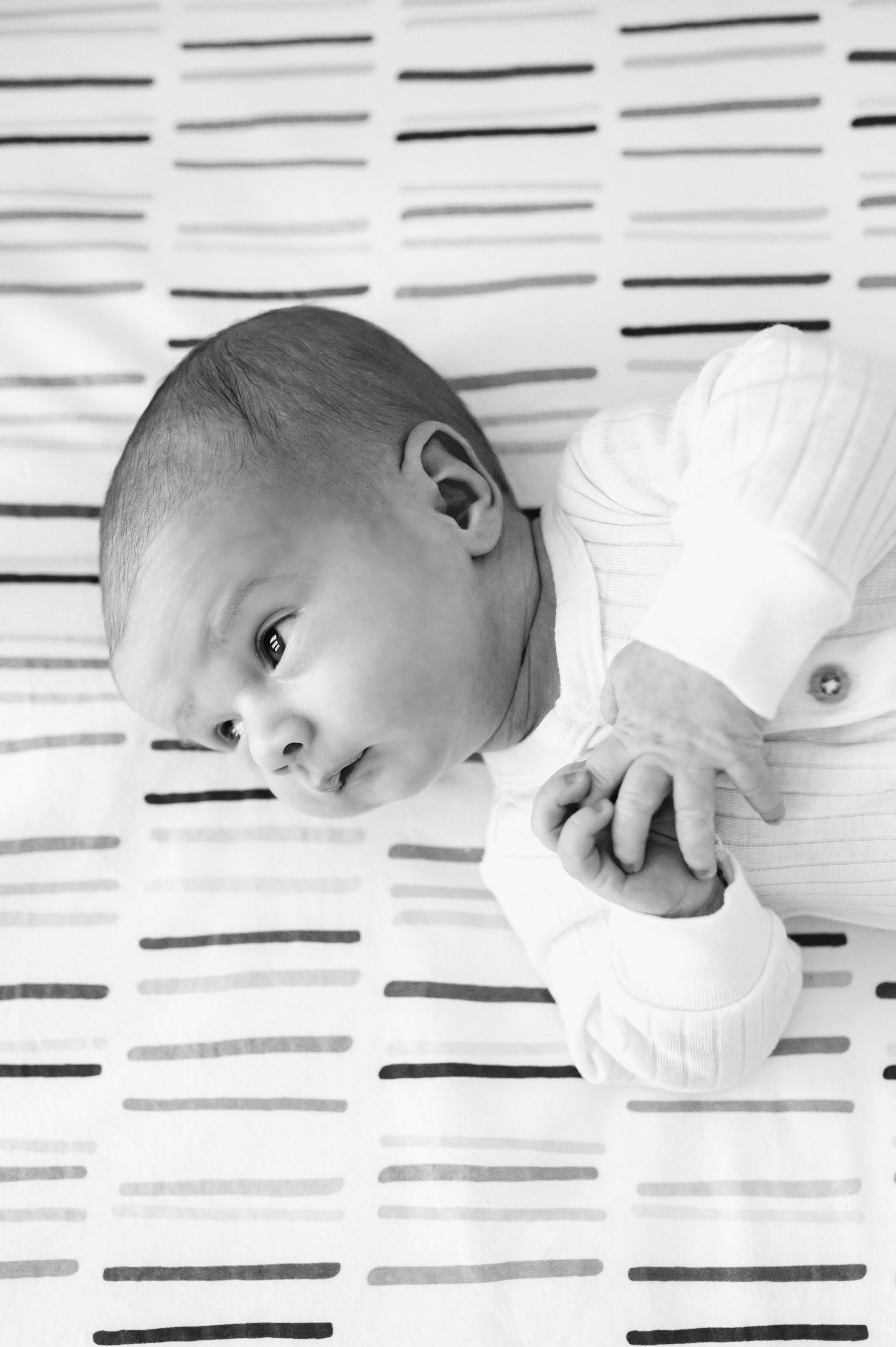 a black and white picture of a baby boy laying on his back on a striped crib sheet and looking over his shoulder toward the light coming in through the window during a newborn photoshoot