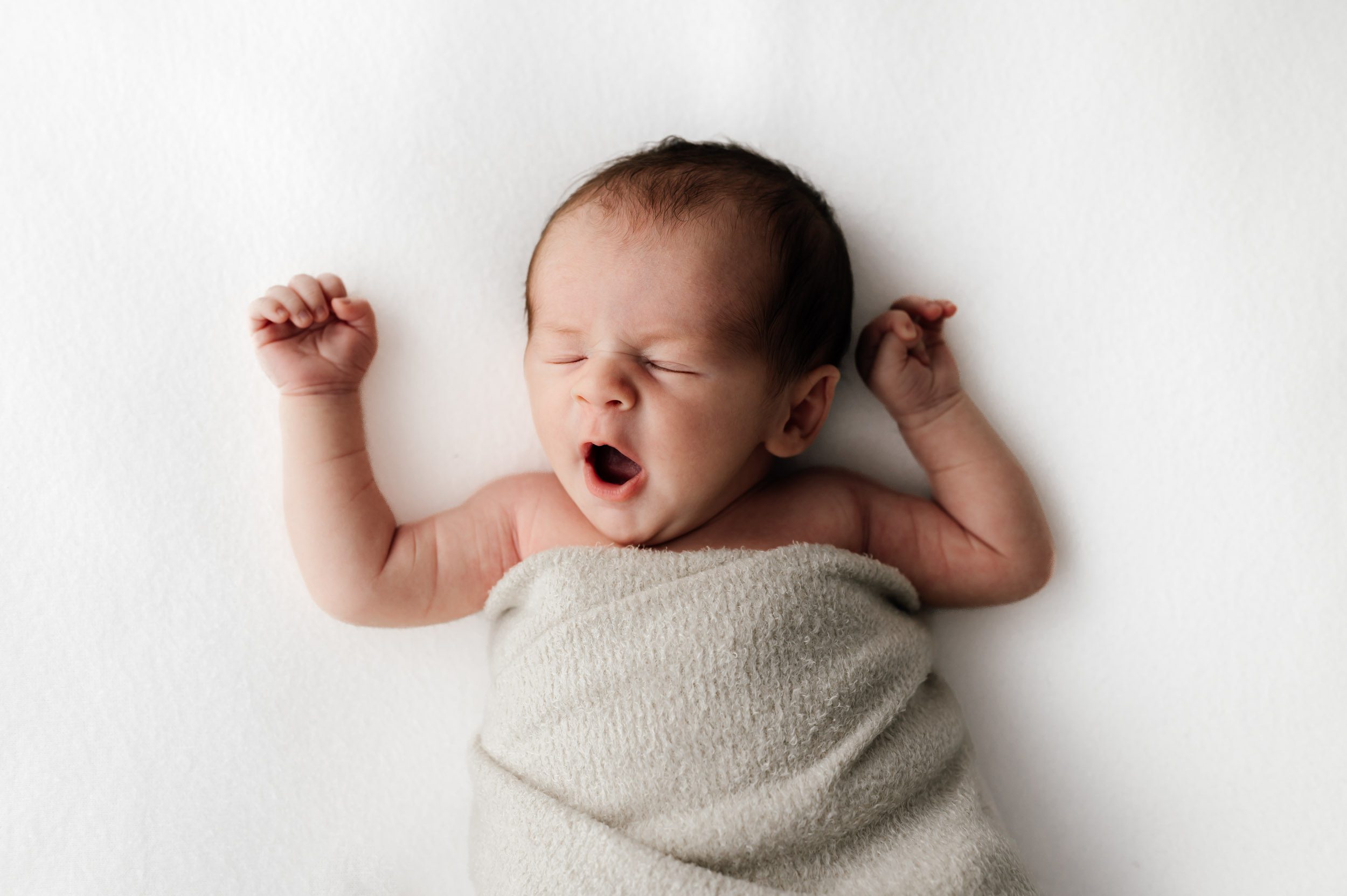 a baby boy laying on a white backdrop and yawning during a newborn photoshoot