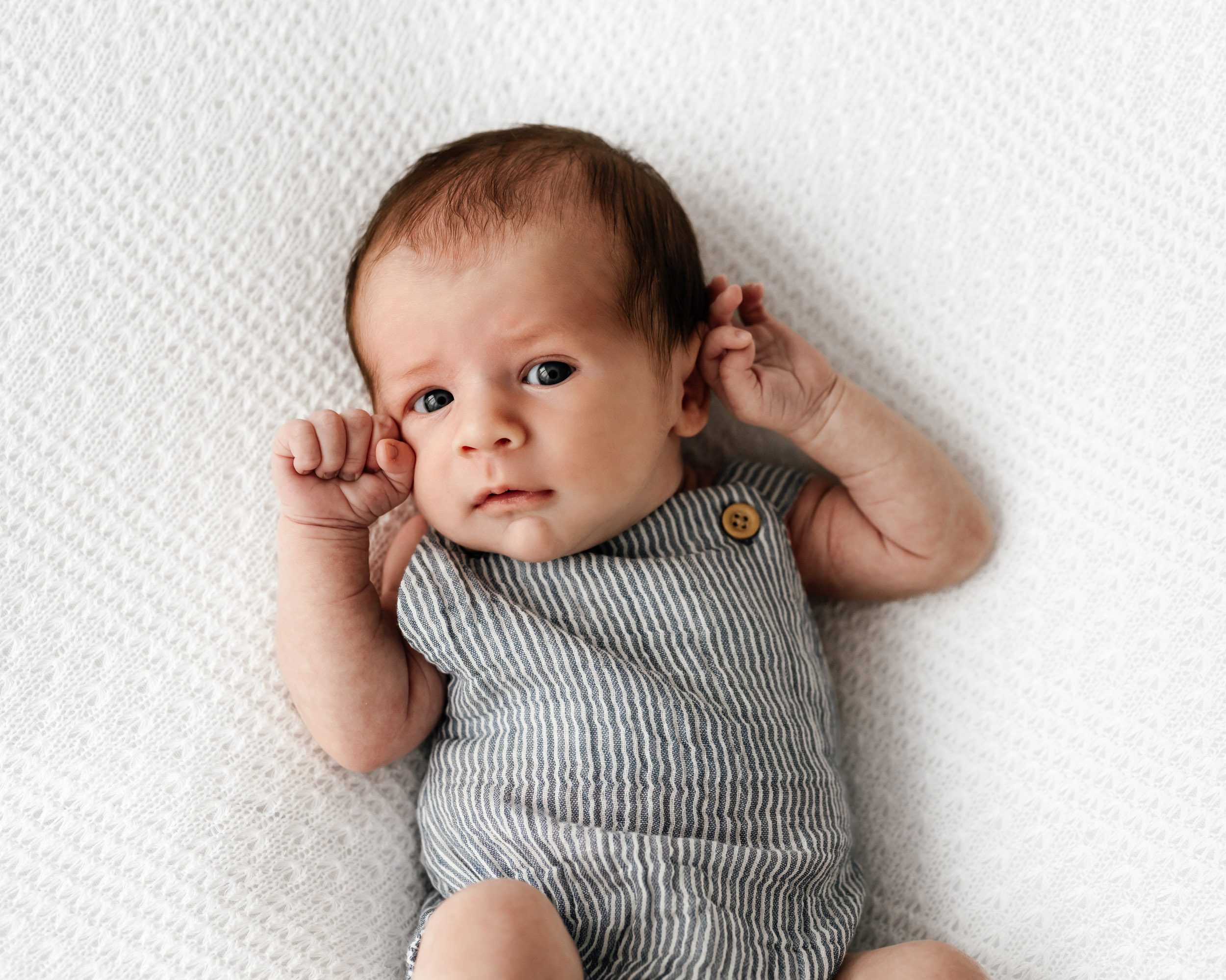 a baby boy laying on a white textured backdrop and holding his hands up to his face while he looks intently at the camera during a newborn photoshoot