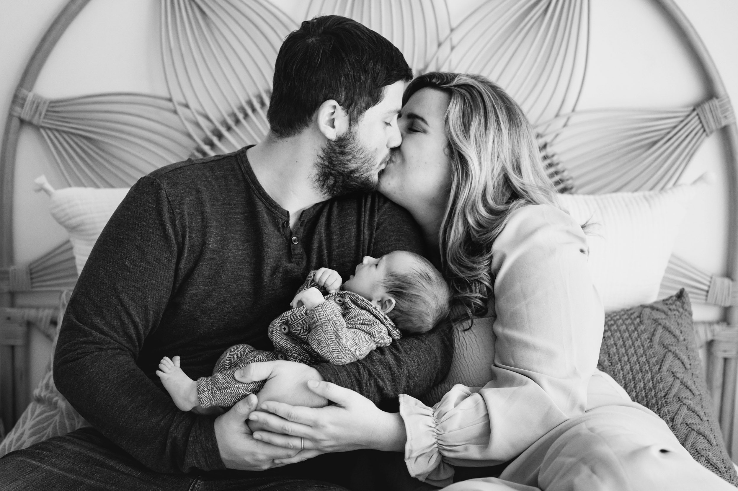 a black and white picture of new parents holding their baby boy and kissing each other during a newborn lifestyle photography session