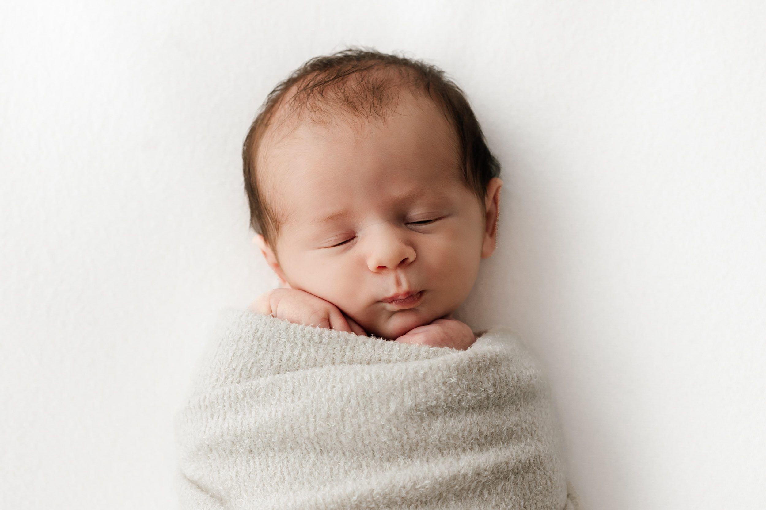 a baby boy wrapped in a fuzzy beige swaddle laying on a a white backdrop and pursing his lips during a newborn photoshoot