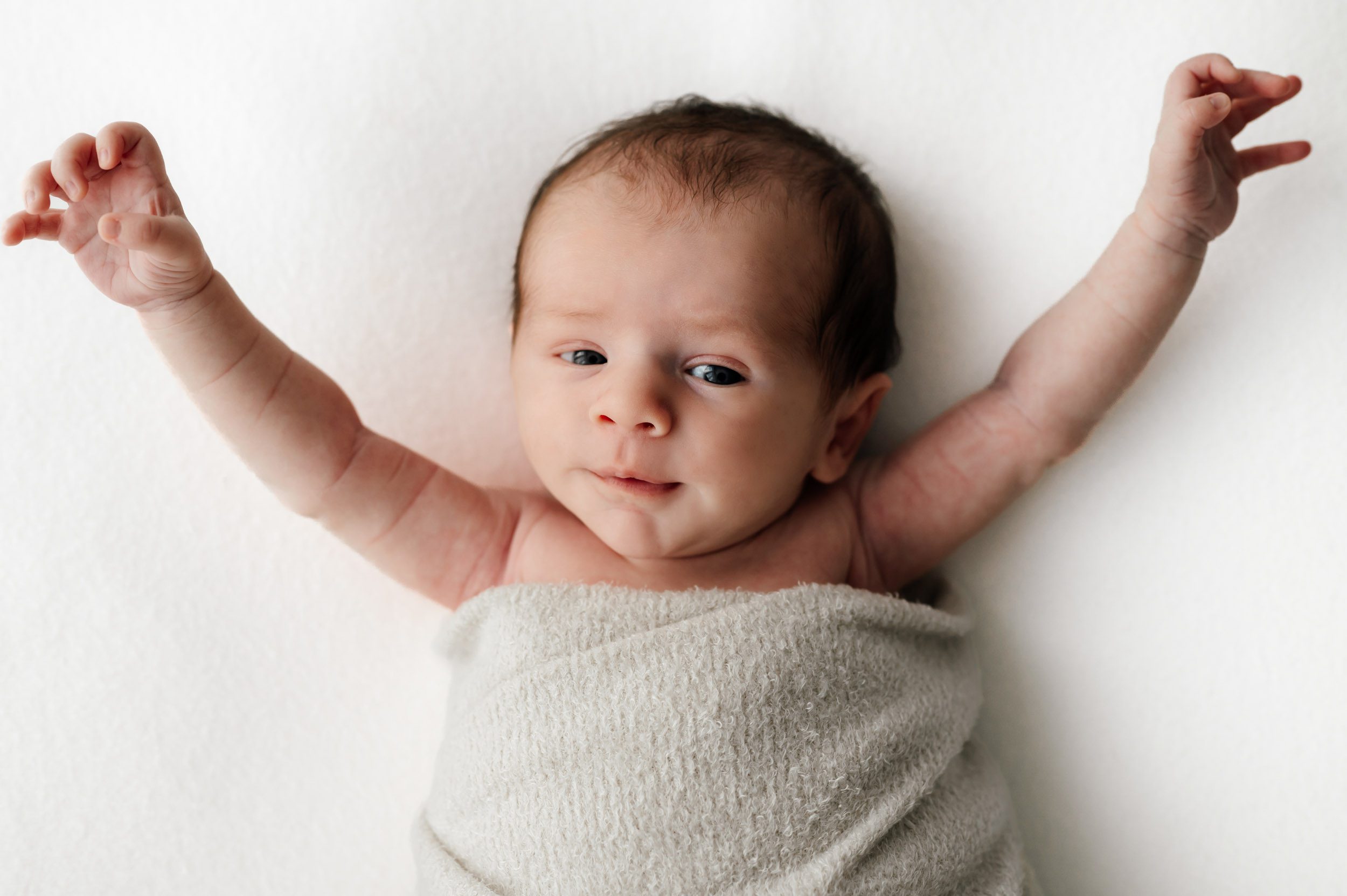 a baby boy laying on a white backdrop and stretching his arms up in the air as he looks toward the camera during a newborn photoshoot