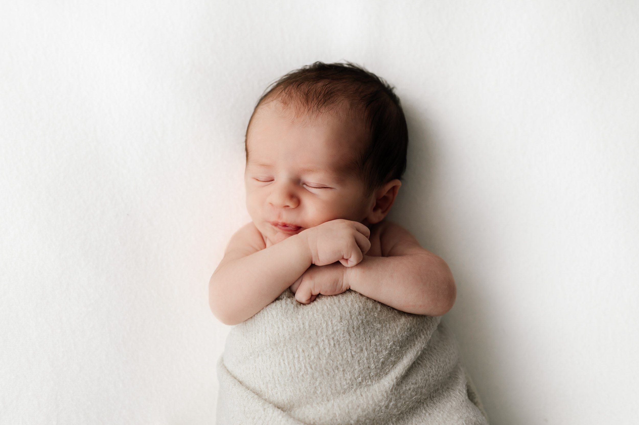 a newborn baby boy laying on a white backdrop and hugging his hands up to his chin with a hint of a smile on his face