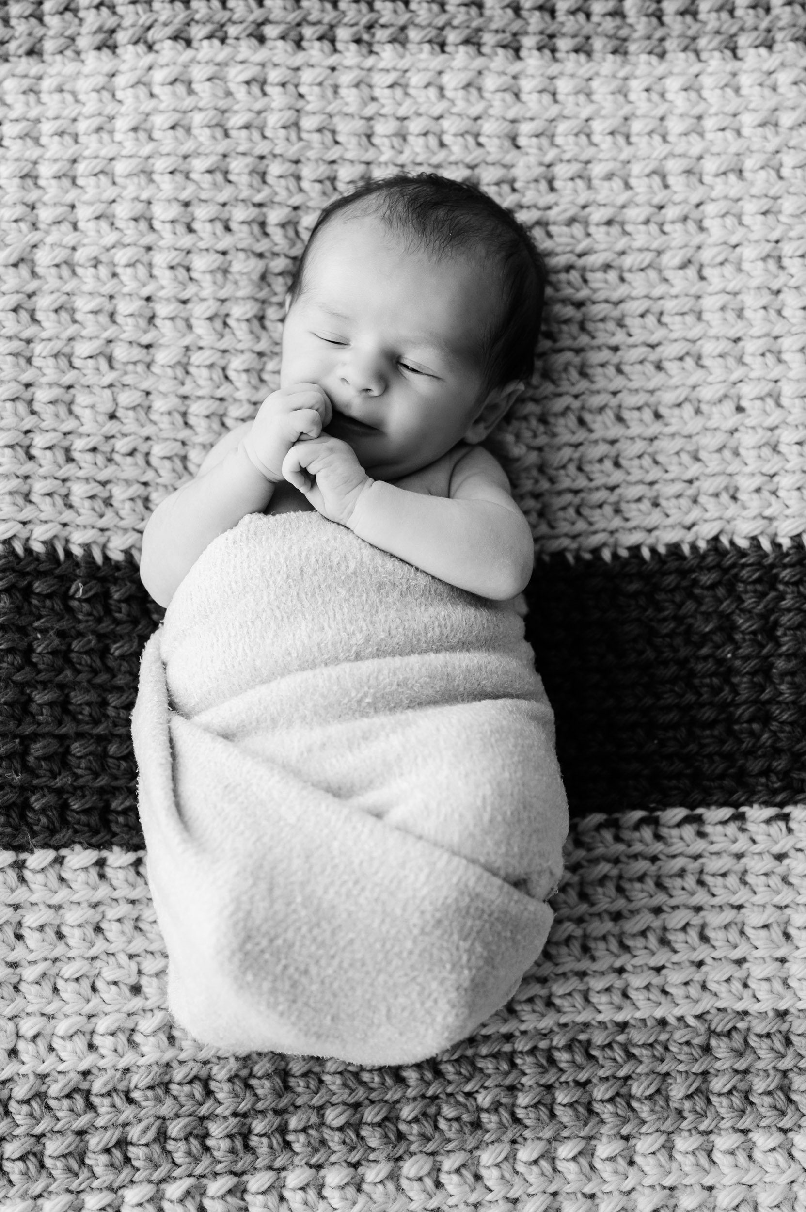 a black and white picture of a baby boy laying on a striped knit blanket with his hands tucked right up against his face during a newborn lifestyle photography session