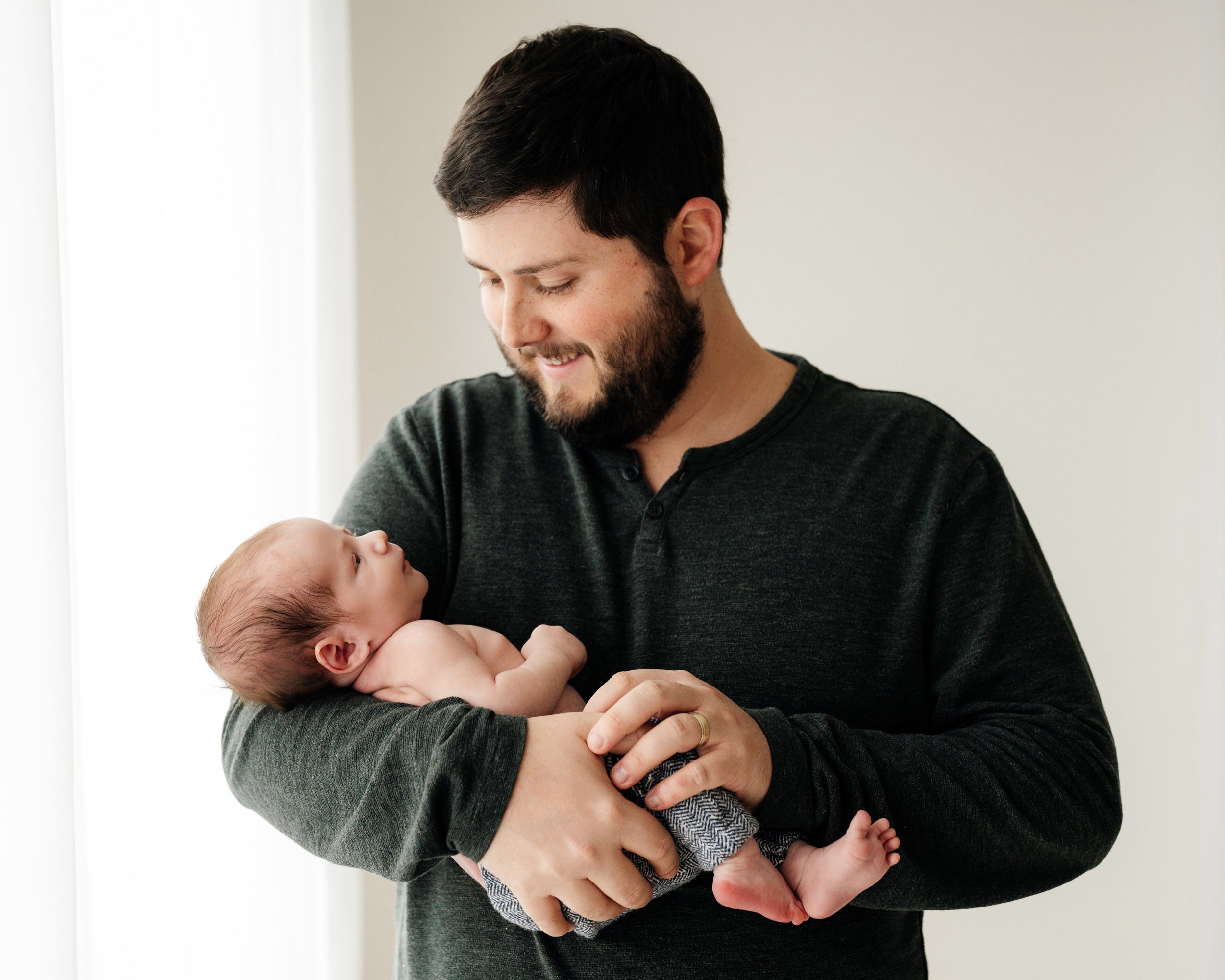 a new father cradling his baby boy in his arms and smiling down at him during a newborn lifestyle photography session