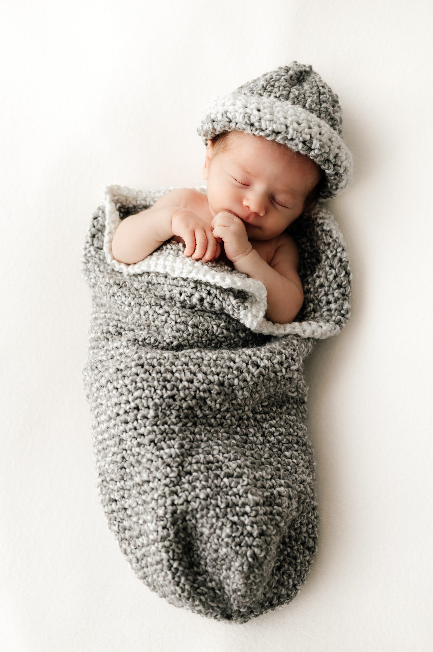 a baby boy laying on a white backdrop in a gray knit hat and sleep sack during a newborn photoshoot