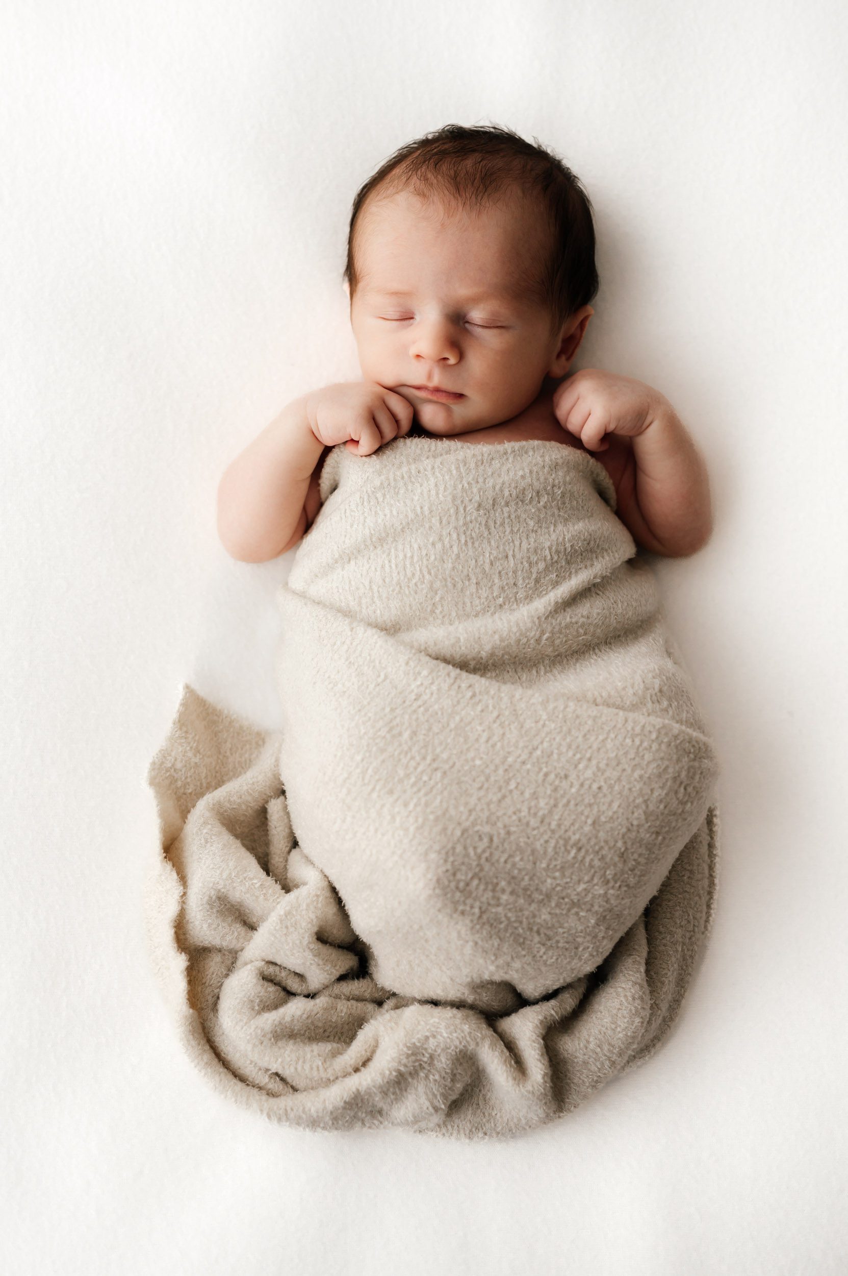 a baby boy wrapped in a beige fuzzy swaddle sleeping on a white backdrop with his hands tucked up against his cheeks during a newborn photo session