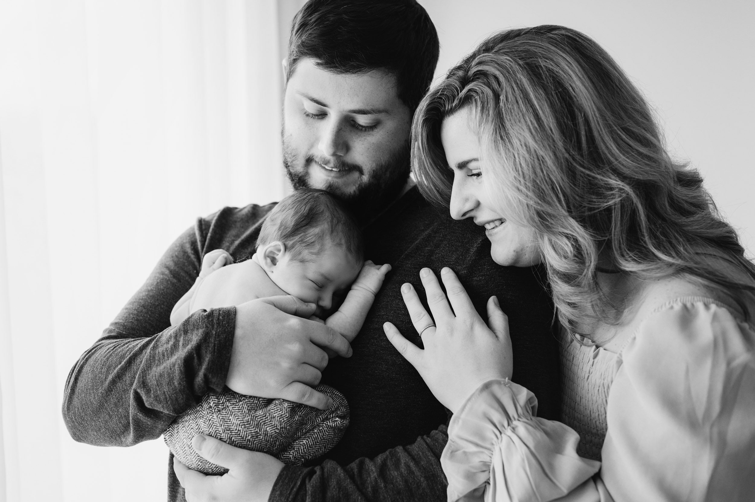 a black and white picture of a new dad hugging his baby boy against his chest while mom rests her head on dad's shoulder and smiles at her baby during a newborn lifestyle photography session