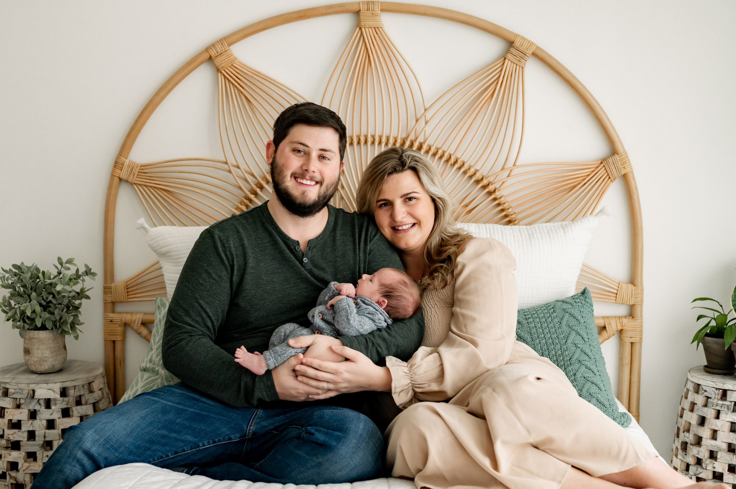 new parents sitting on a bed holding their baby boy in their arms and smiling at the camera during a newborn lifestyle photography session