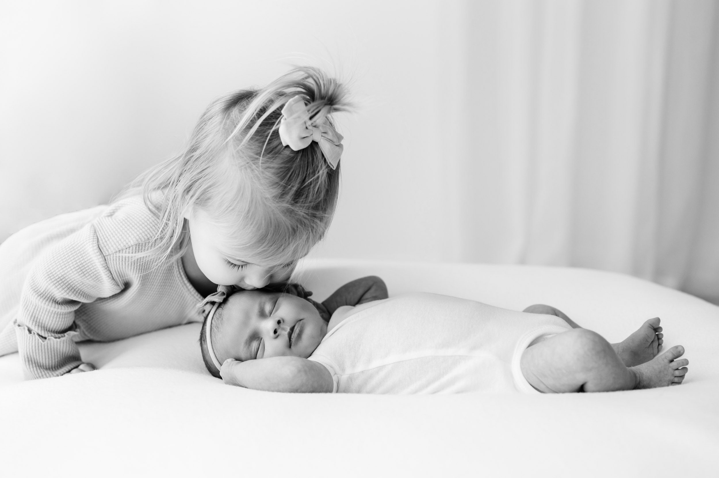 a black and white picture of a baby girl laying on a white bean bag while her older sister gently kisses her on the head during a newborn photo session