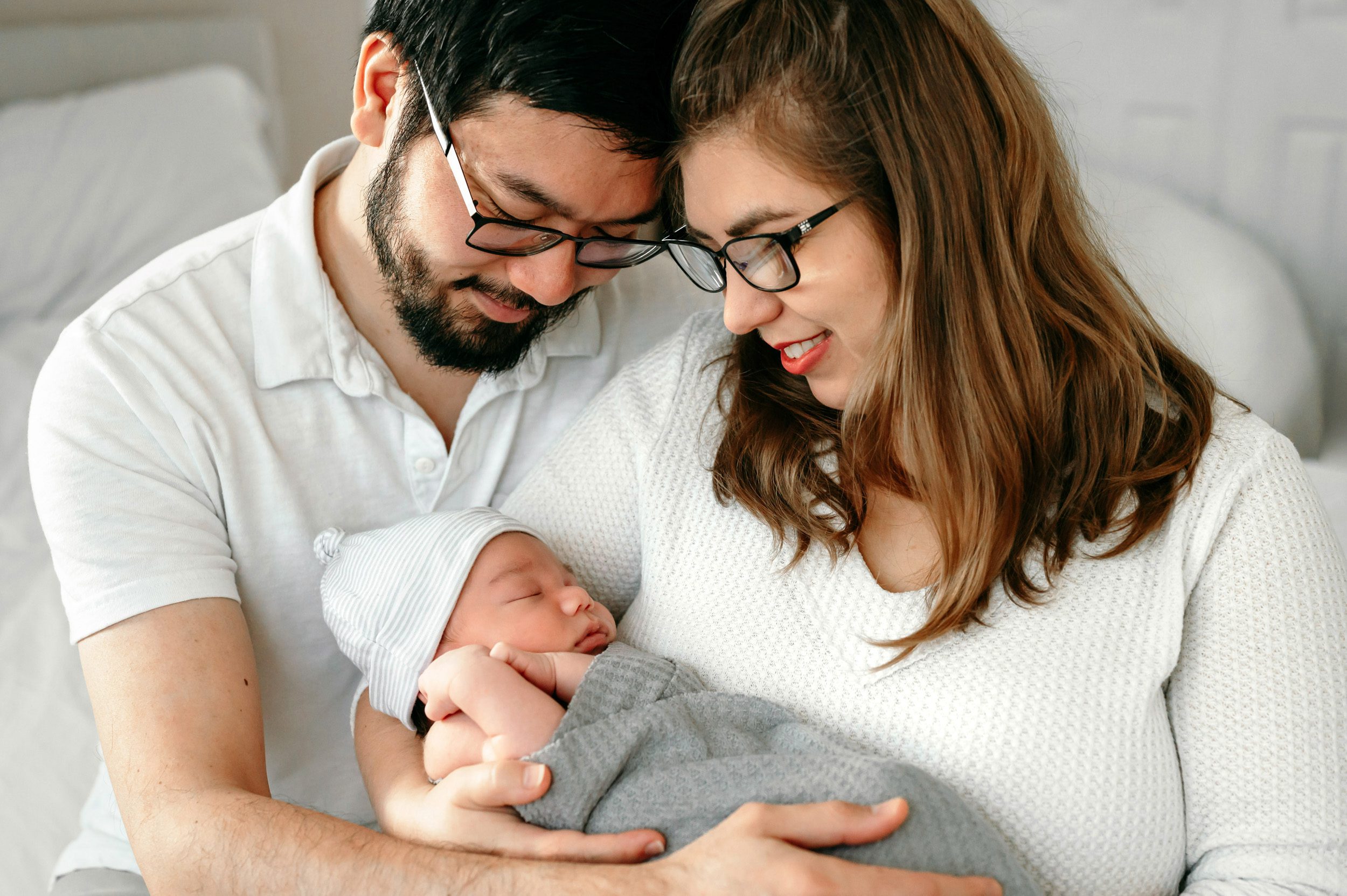 new parents wearing white shirts holding their baby boy in their arms and resting their foreheads together as they gaze down at him adoringly during a newborn photos session