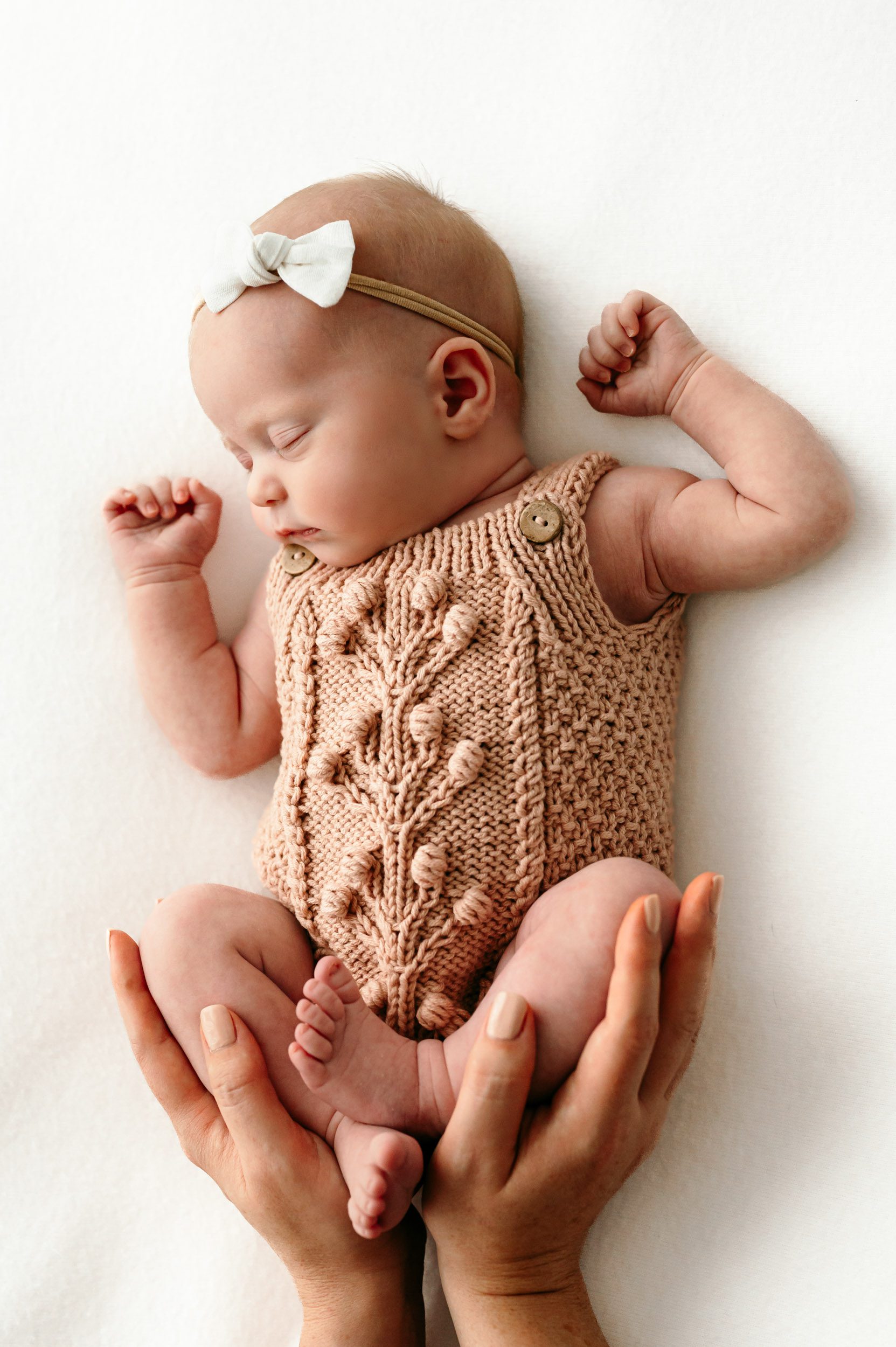 a baby girl wearing a pink knit onesie and a white headband sleeping on a white backdrop while mom cradles her legs in her hands during a newborn photos session