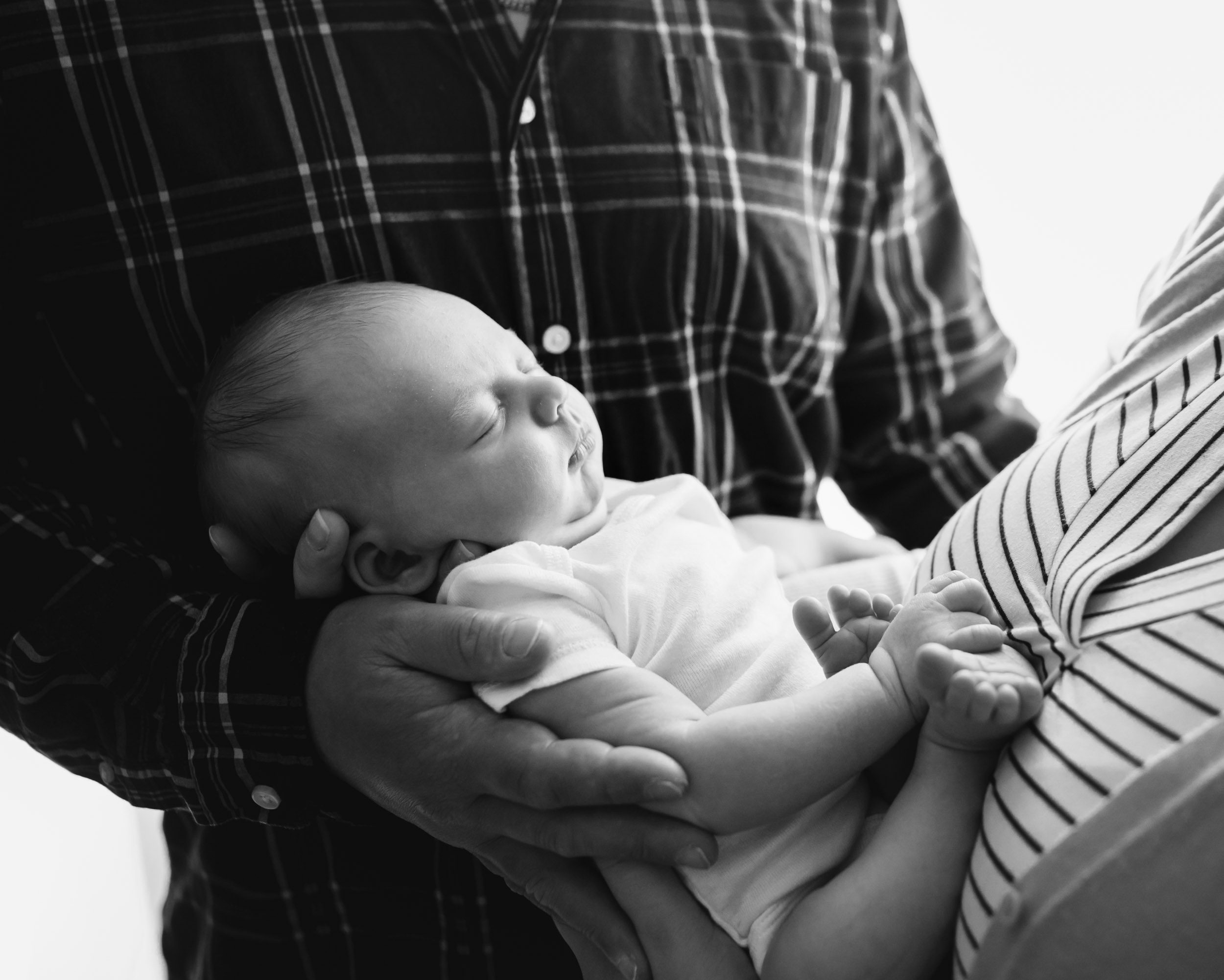 a black and white close up picture of a baby boy snugged in his mom's arms with his tiny feet tucked up against her chest while dad cradles him in his hand during a newborn photo session