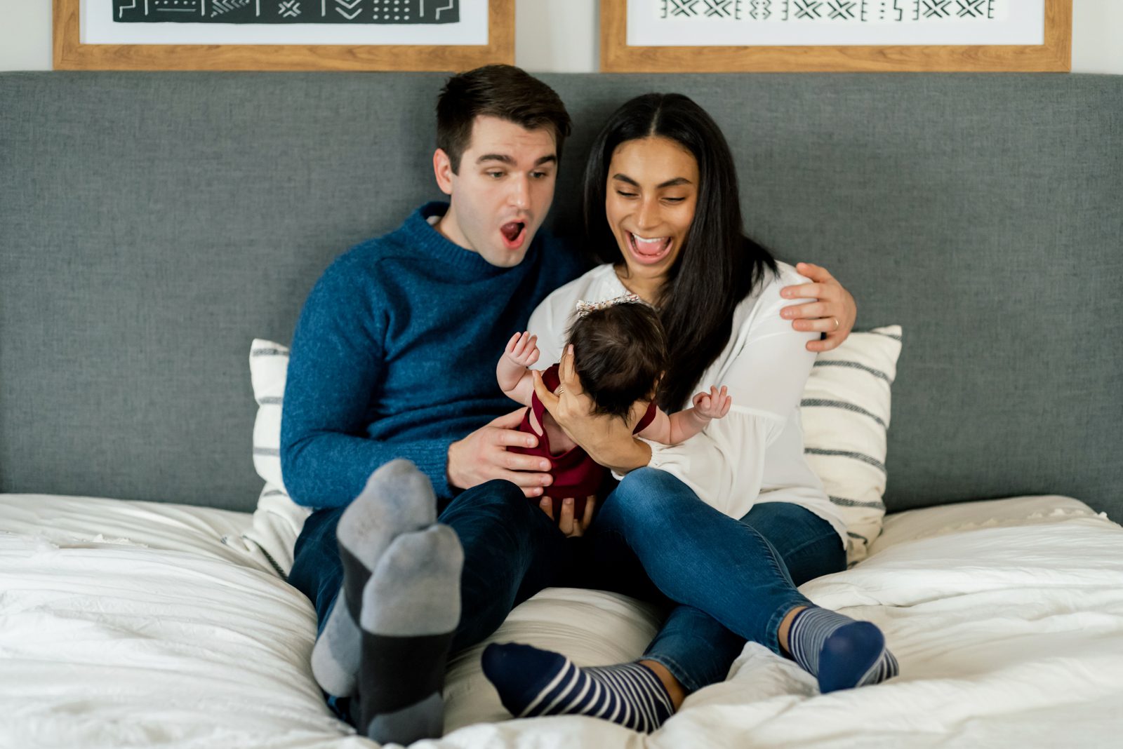 a mom and dad sitting on their bed holding their baby girl in their arms and making silly faces at her during a newborn photo session