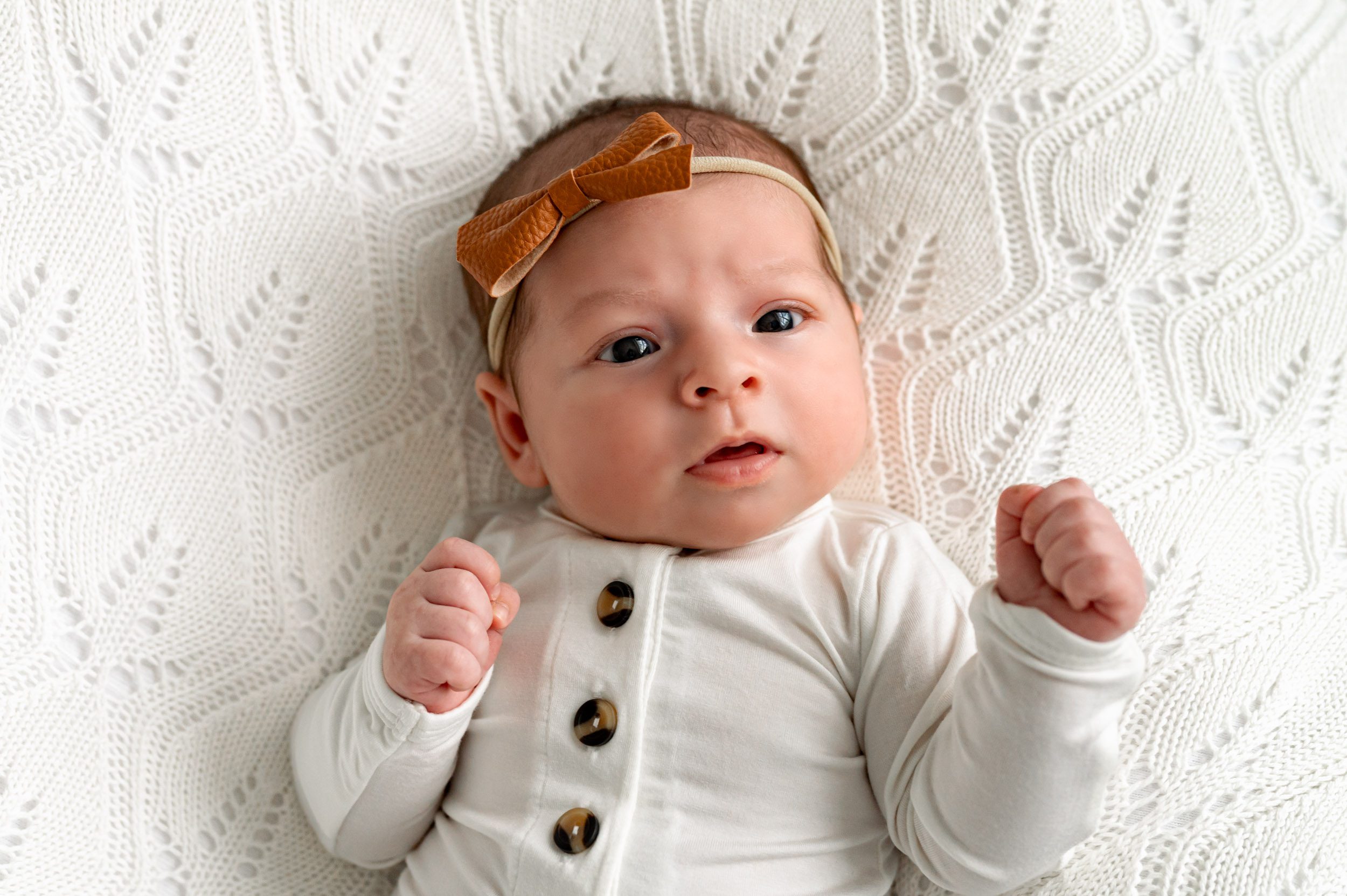 a baby girl wearing a long sleeve white onesie with multicolored buttons and a rust colored leather headband laying on a white backdrop and gazing up toward the camera with wide open eyes during a newborn photos session